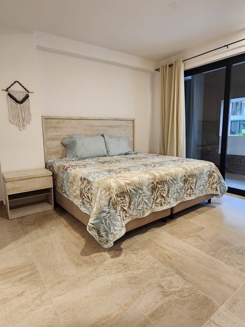 Bedroom with a wooden bedframe, tropical-patterned bedding, light curtains, and beige tiled floor.