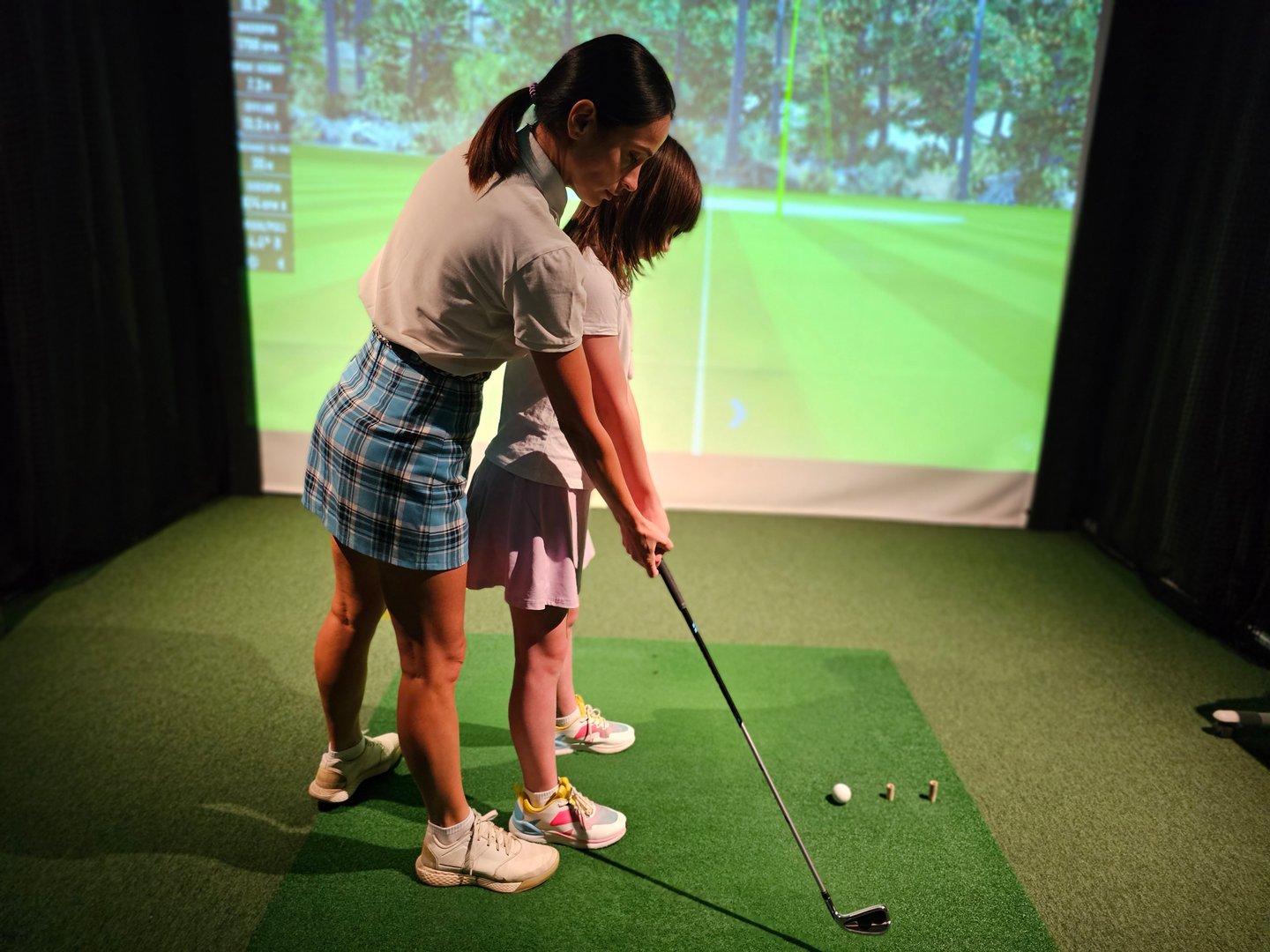 Golf training session with an instructor guiding a young learner indoors with a virtual simulation backdrop
