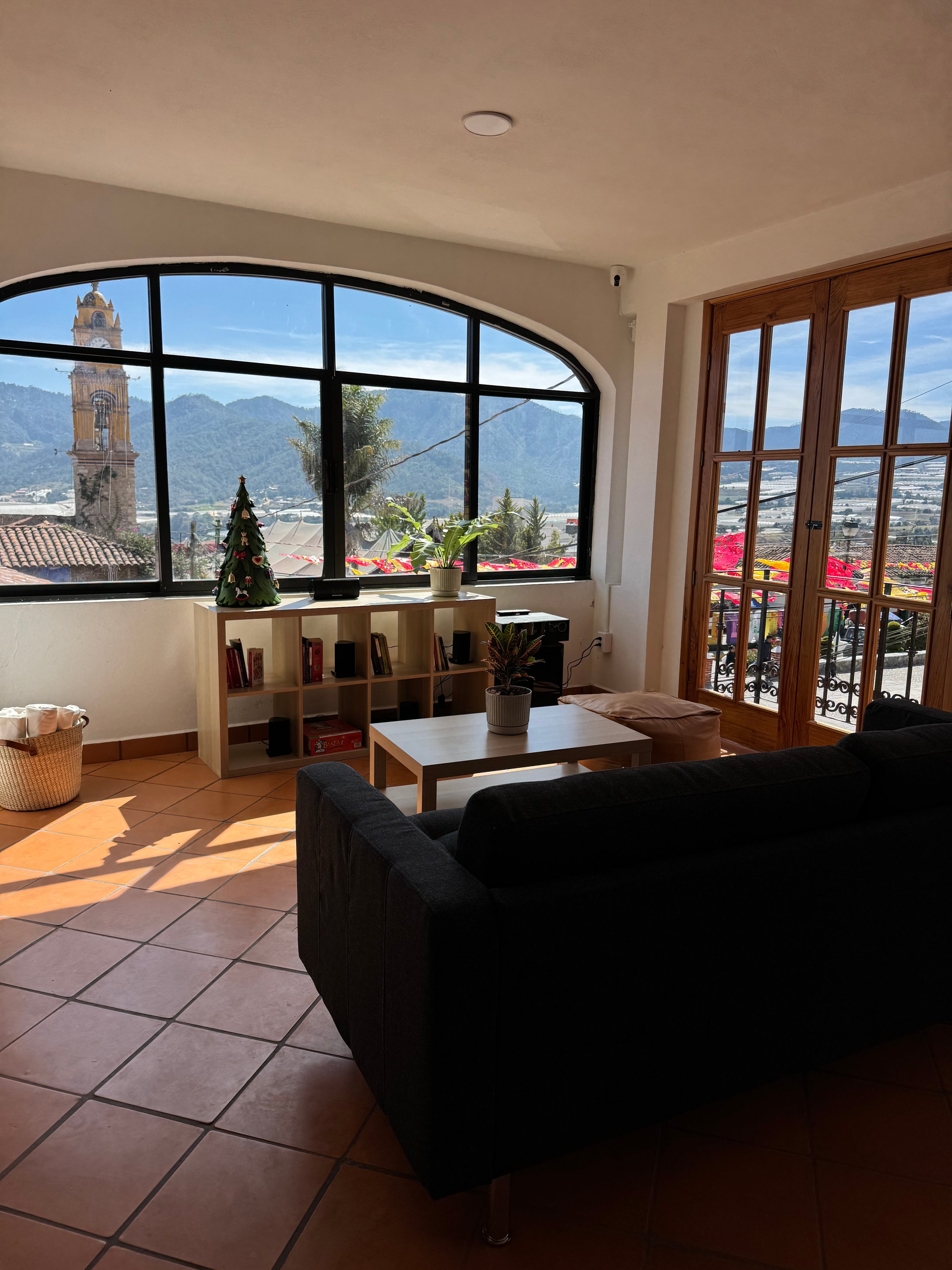 Sunlit room with a couch, small table, bookshelf, and Christmas tree, offering a view of a bell tower and mountains.