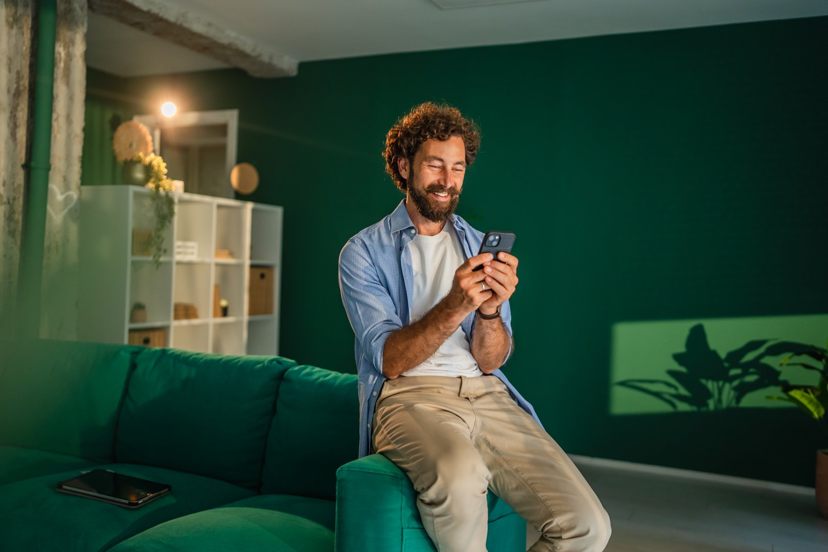 Smiling young businessman using mobile phone app while sitting on a comfortable sofa in his start up company office. Enjoying success. Reading good news. Chatting with friends or browsing social media