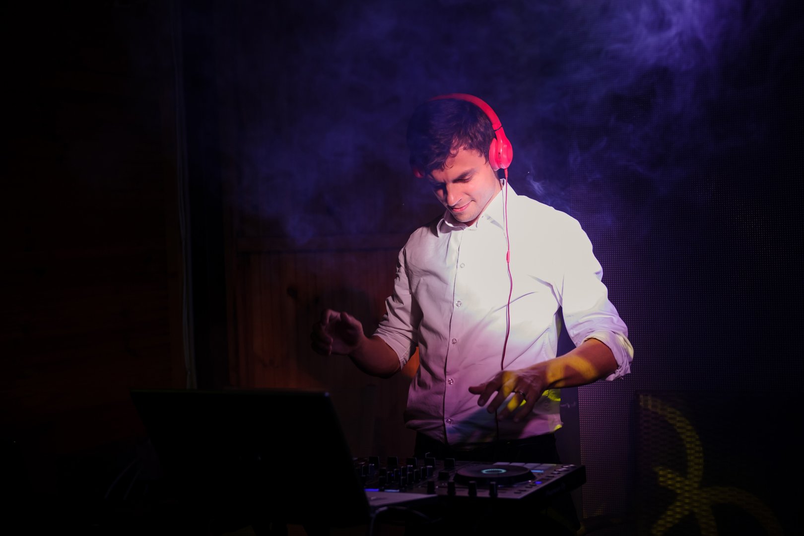 Young and beautiful man DJ/ disc jockey plays and mixing on CD players/dj mixer/mixing console at nightclub during party/ wedding party .EDM. Black background with light and smoke