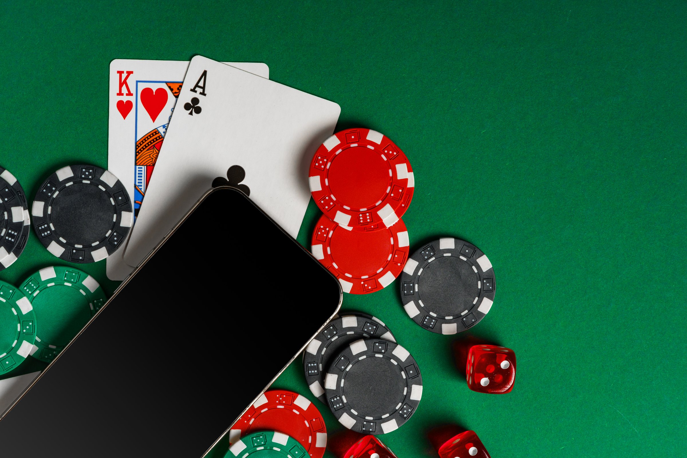 This image shows a smartphone with a blank screen lying on a green felt table surface surrounded by playing cards, chips, and dice.