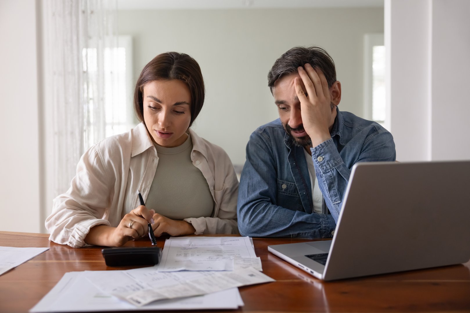 Concerned young middle aged couple counting expenses, finding overspending, money loss, risk of bankruptcy, using calculator at table with paper bills and laptop, touching head