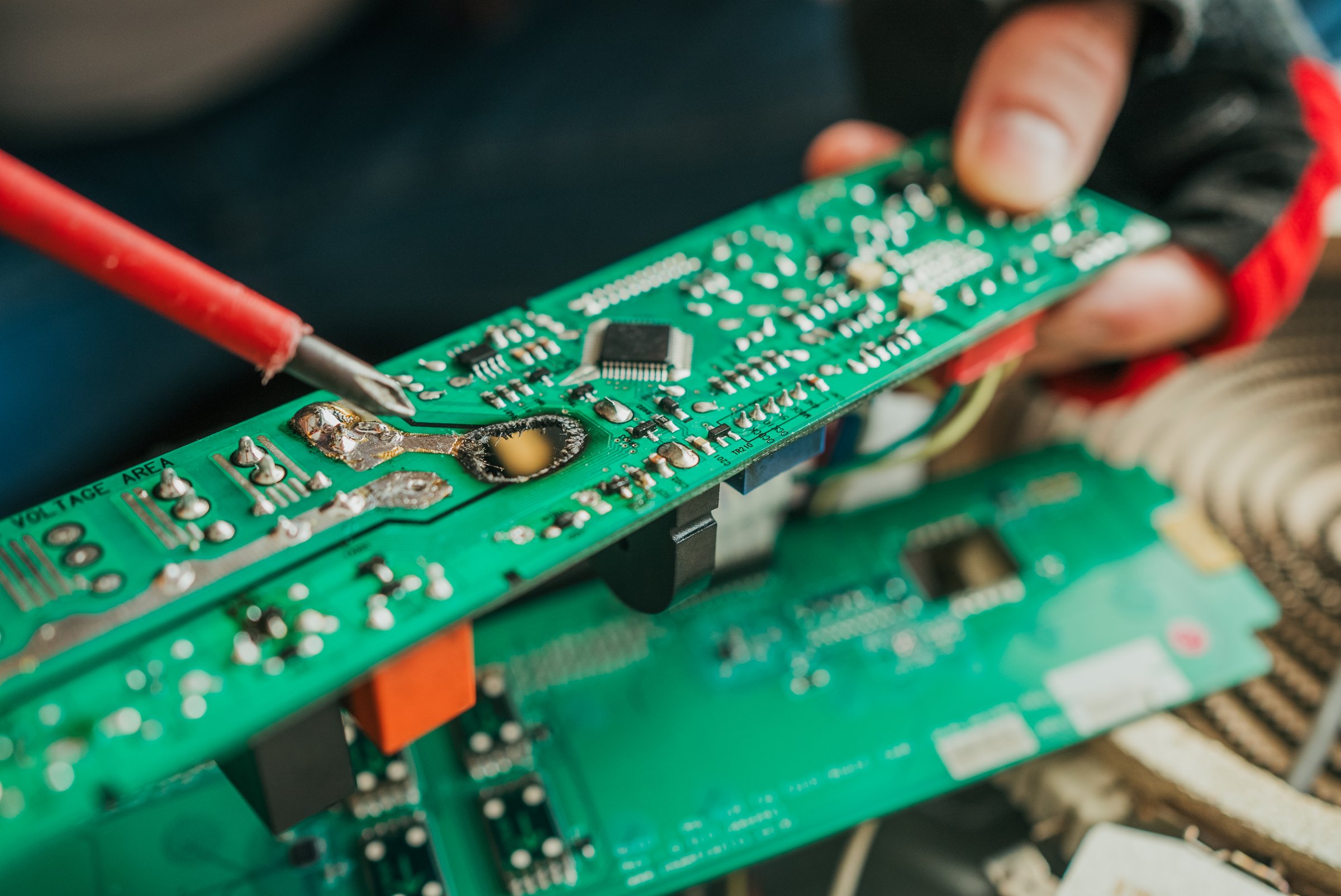 Close-up of a technician using a soldering iron to repair a burnt and damaged electronic circuit board, showcasing the intricate components and the precision required in electronics repair