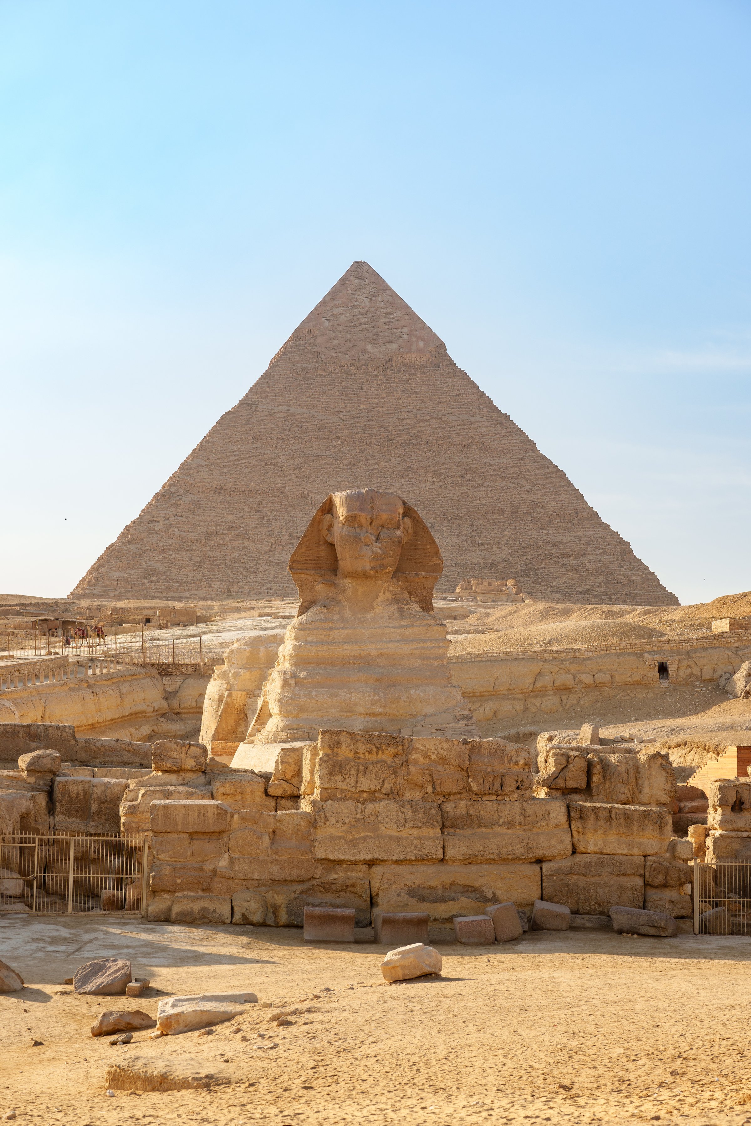 A majestic Sphinx rests in the foreground, overlooking the iconic Great Pyramid of Giza on a sunny afternoon. Tourists explore the ancient site nearby, marveling at the history.