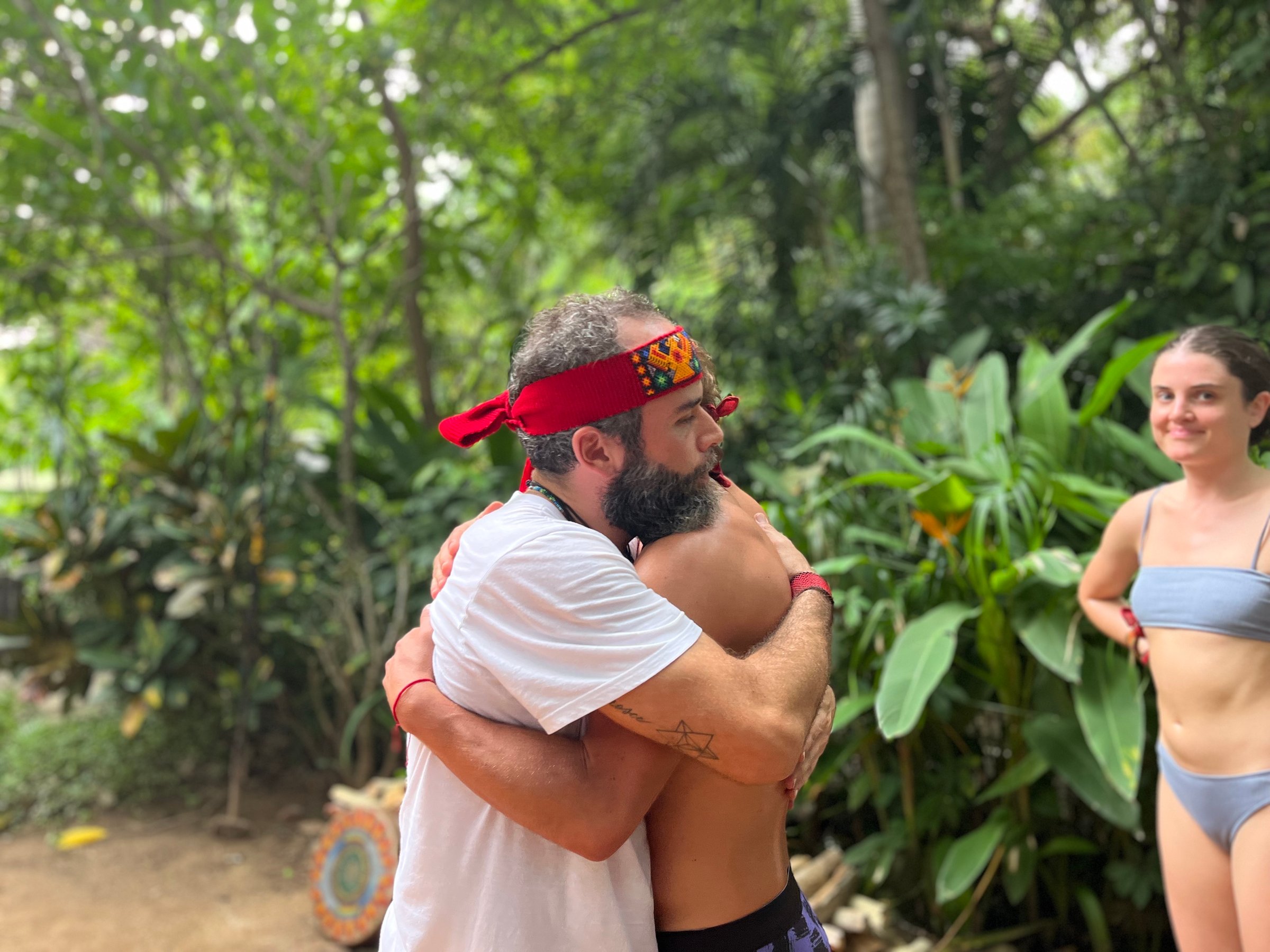 Two people hugging in a lush garden, wearing casual clothes and red headbands. Another person stands nearby, smiling.