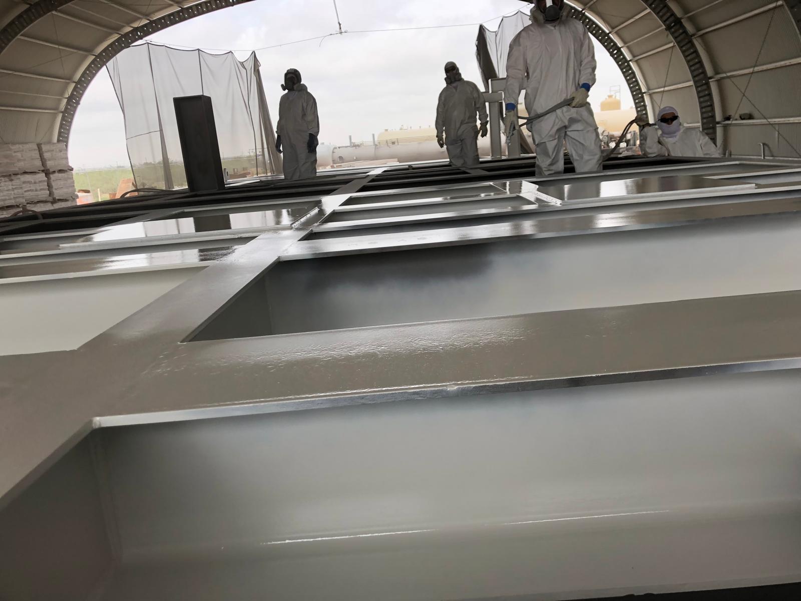 Workers in protective suits applying a coating to large metal structures inside a covered industrial facility.