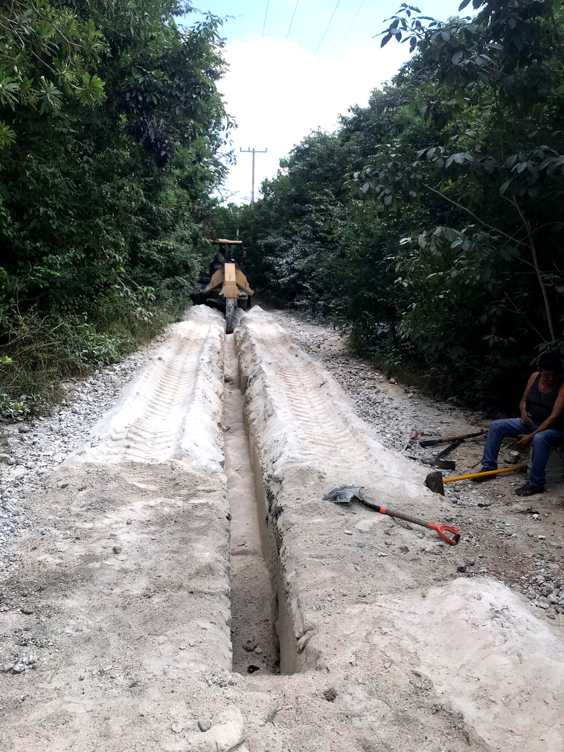 A narrow trench being dug along a dirt road in a wooded area, with heavy machinery and a worker resting nearby.