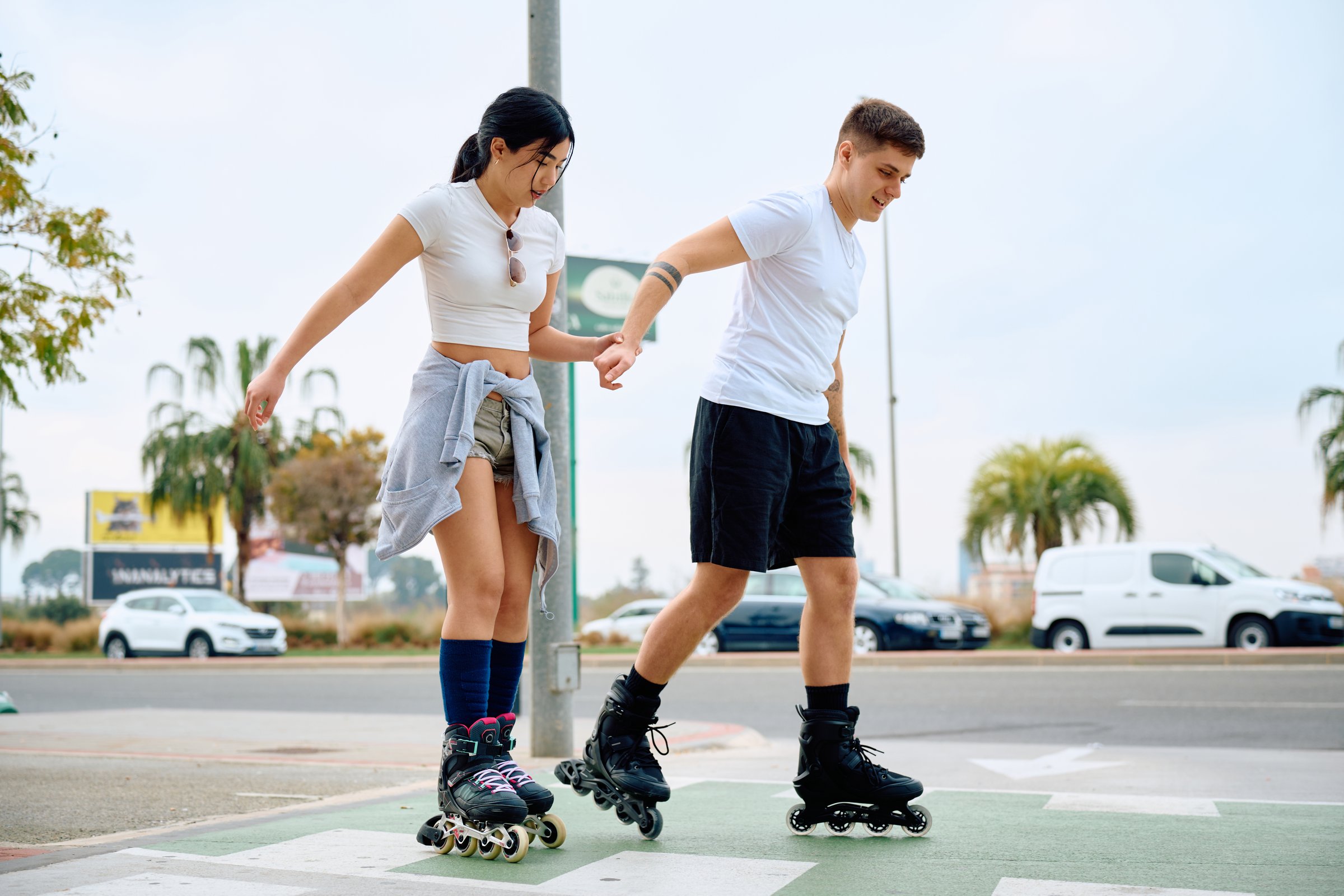 A young couple enjoys roller skating hand in hand on a smooth pavement in a lively urban environment. The sun shines brightly, highlighting their playful interaction.