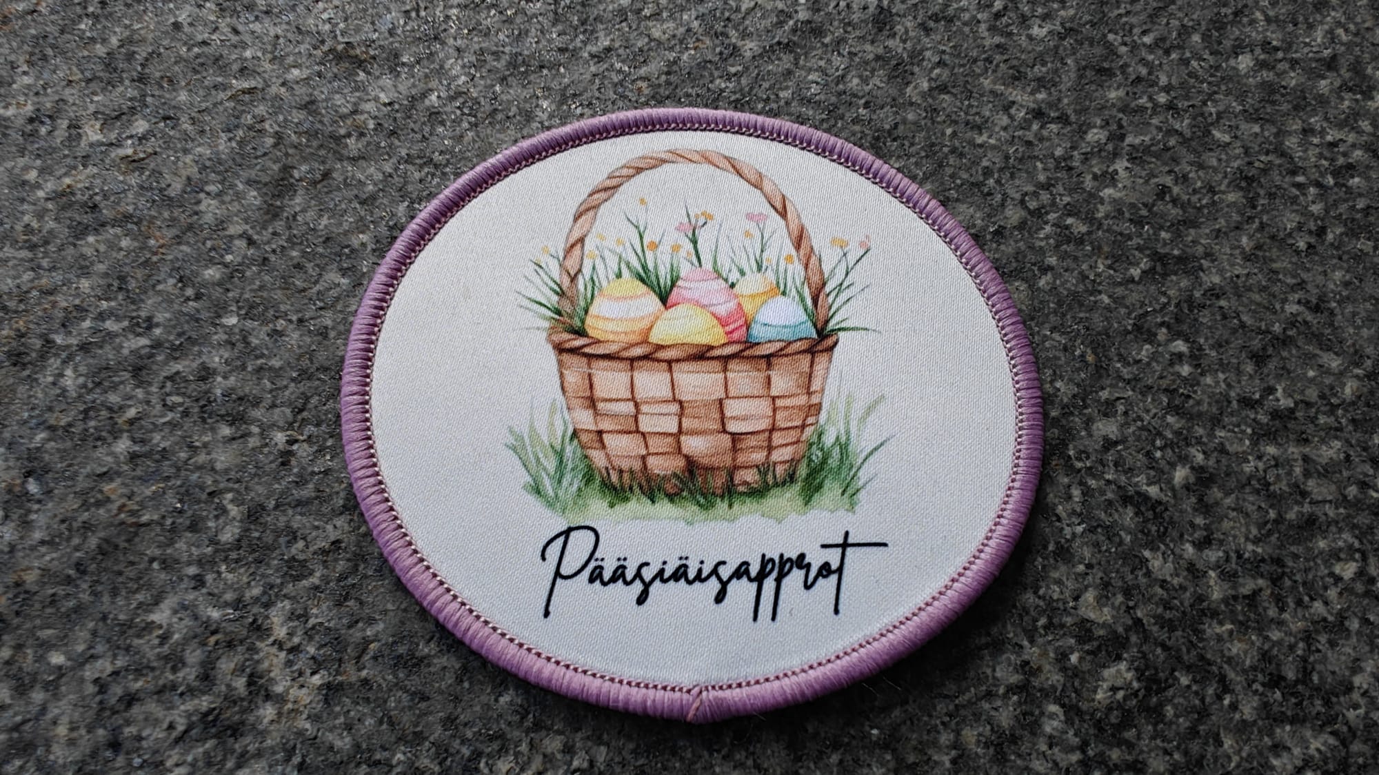 Round embroidered patch with a basket of Easter eggs and flowers, featuring the text Pääsiäisapplot on a stone background.