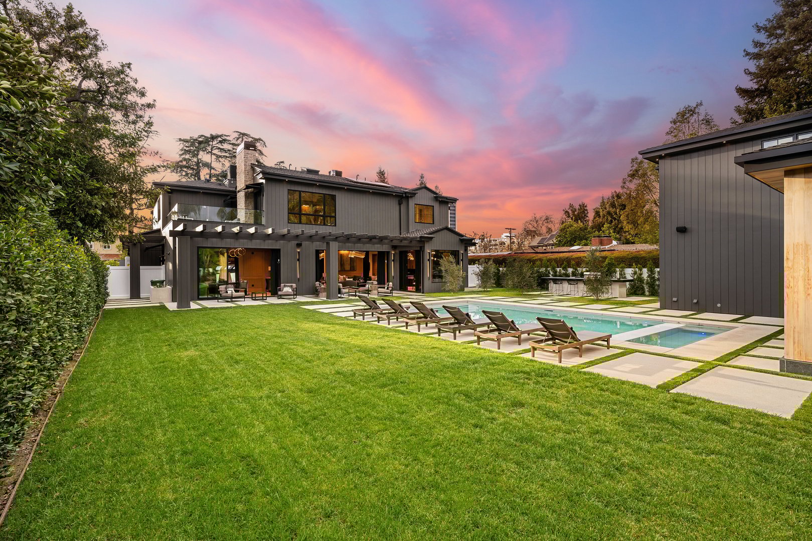 A luxurious modern house with a large backyard, swimming pool, and lush green lawn at sunset.