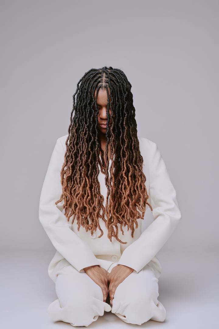 Woman with long dreadlocks sitting down, hands resting on knees, eyes closed in meditation, wearing white suit and peaceful expression on face