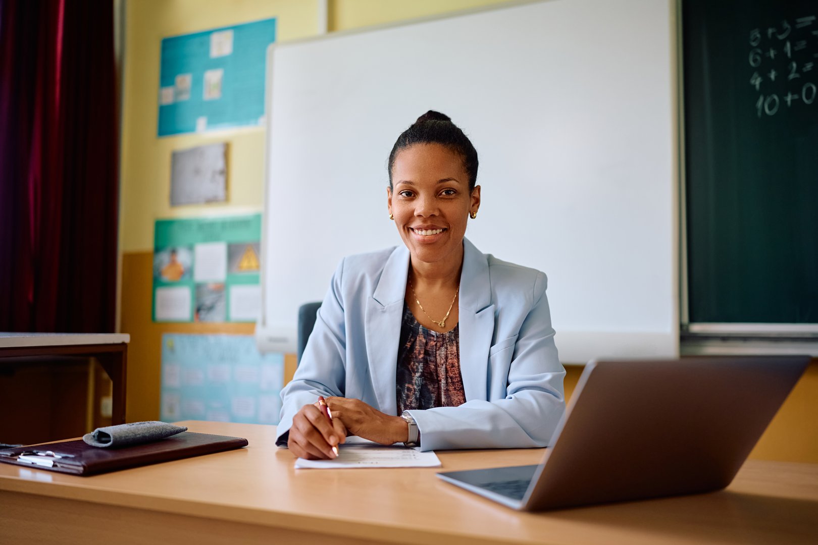 Happy African American elementary school teacher at her desk in the classroom looking at camera.