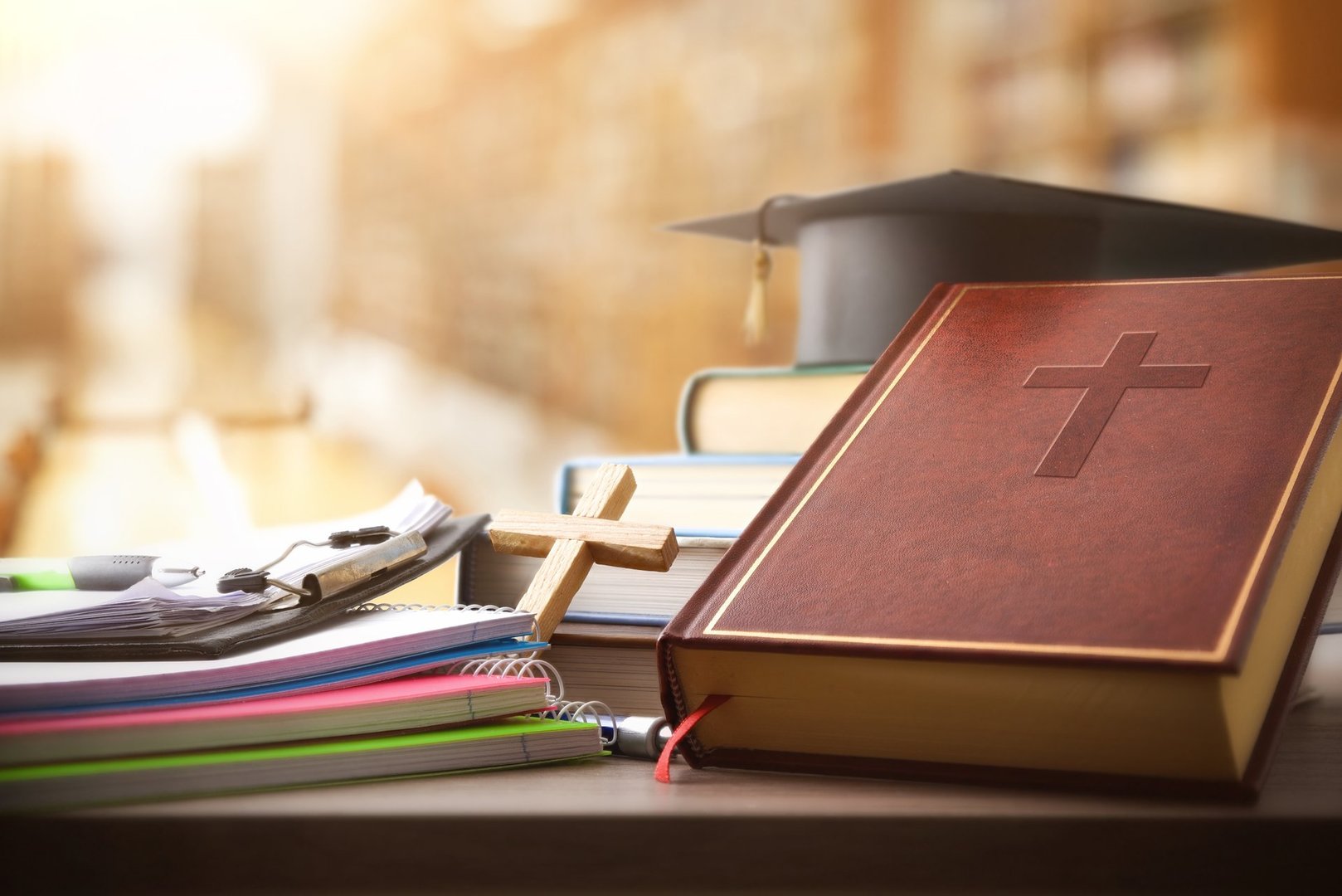 Bible, books, school supplies, and a cap for Catholic education on a wooden table in a library with shelves full of books in the background. Front view.