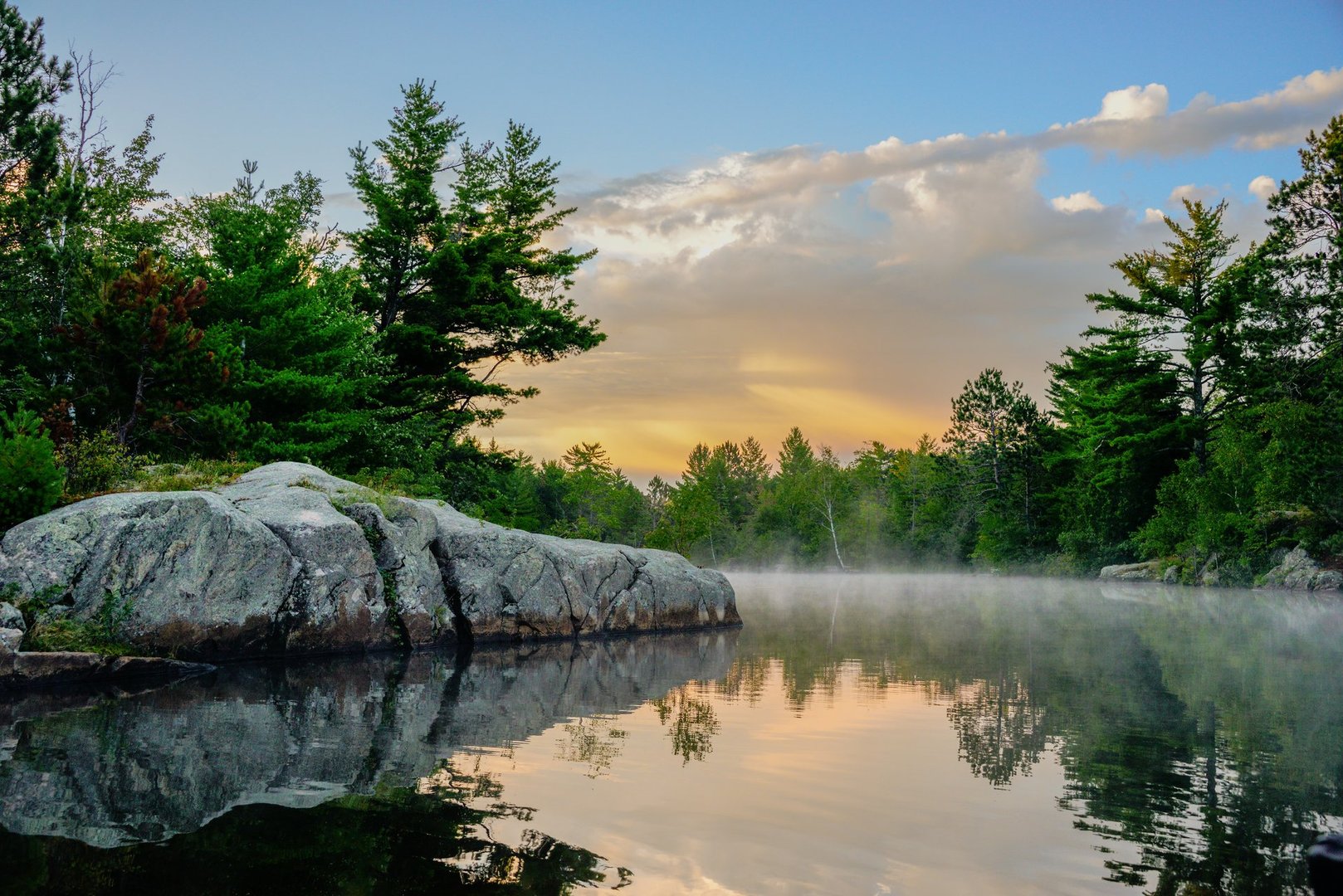 Peaceful morning on the water. Sun and mist reflecting in the lake. Trees and rock boulder on shoreline. Minnesota summer