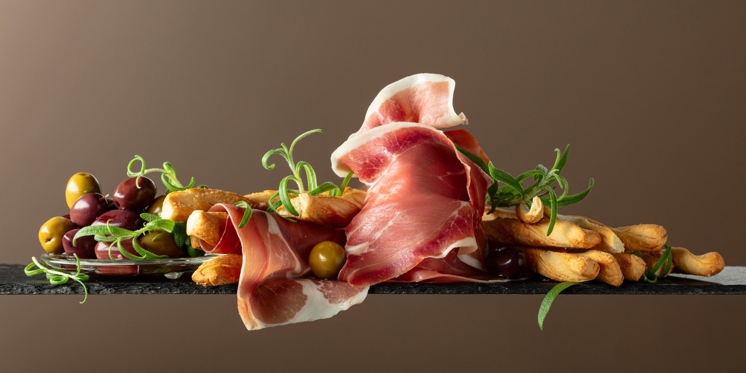 Prosciutto with bread sticks, olives, and rosemary. Traditional Italian snacks on a brown background.