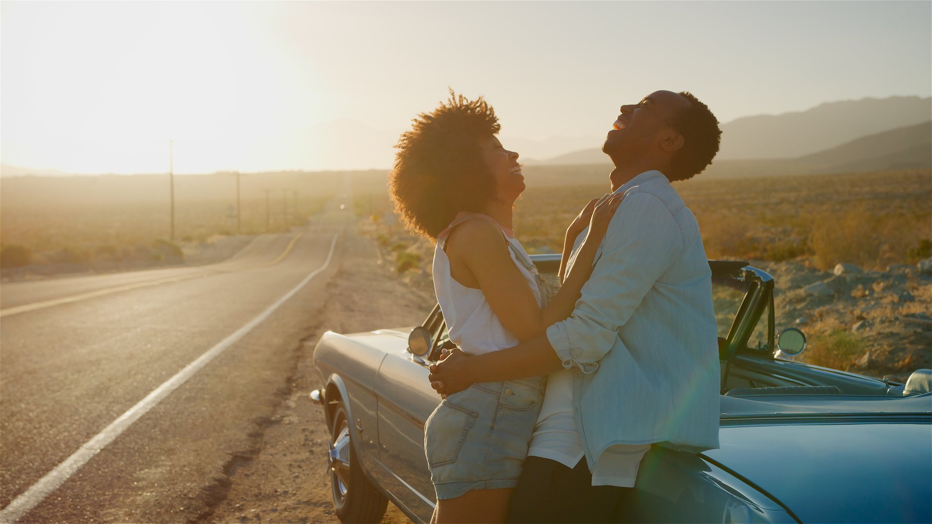 Loving Couple Next To Classic Open Top Car On Road Trip Through Countryside At Sunset