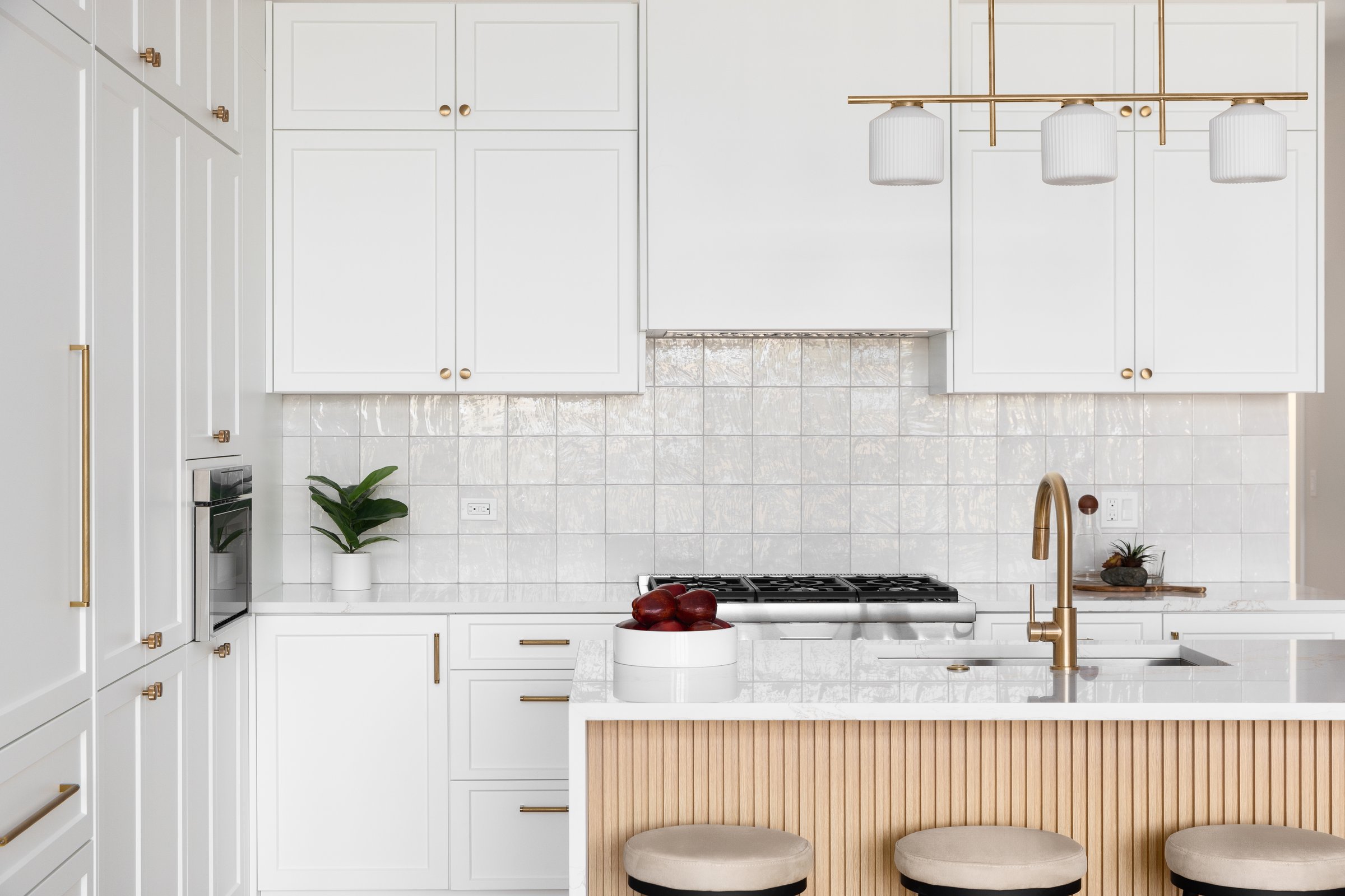 A kitchen detail with white cabinets and square tile backsplash, a waterfall marble countertop and wood slat panel front, and a gold light fixture, hardware, and faucet. No brands or labels.