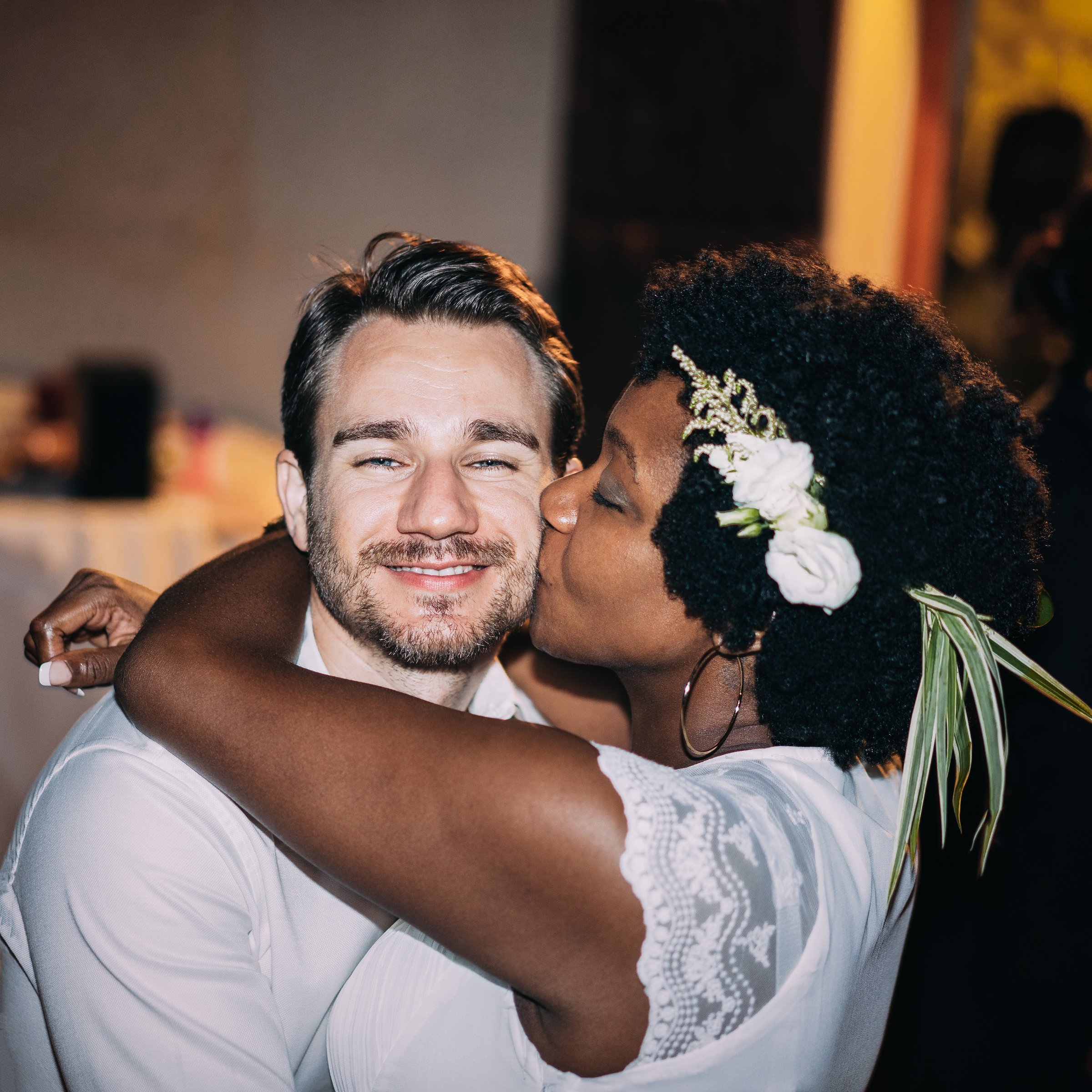 A woman with a floral hair accessory kisses a smiling man on the cheek, both wearing white attire, embracing warmly.