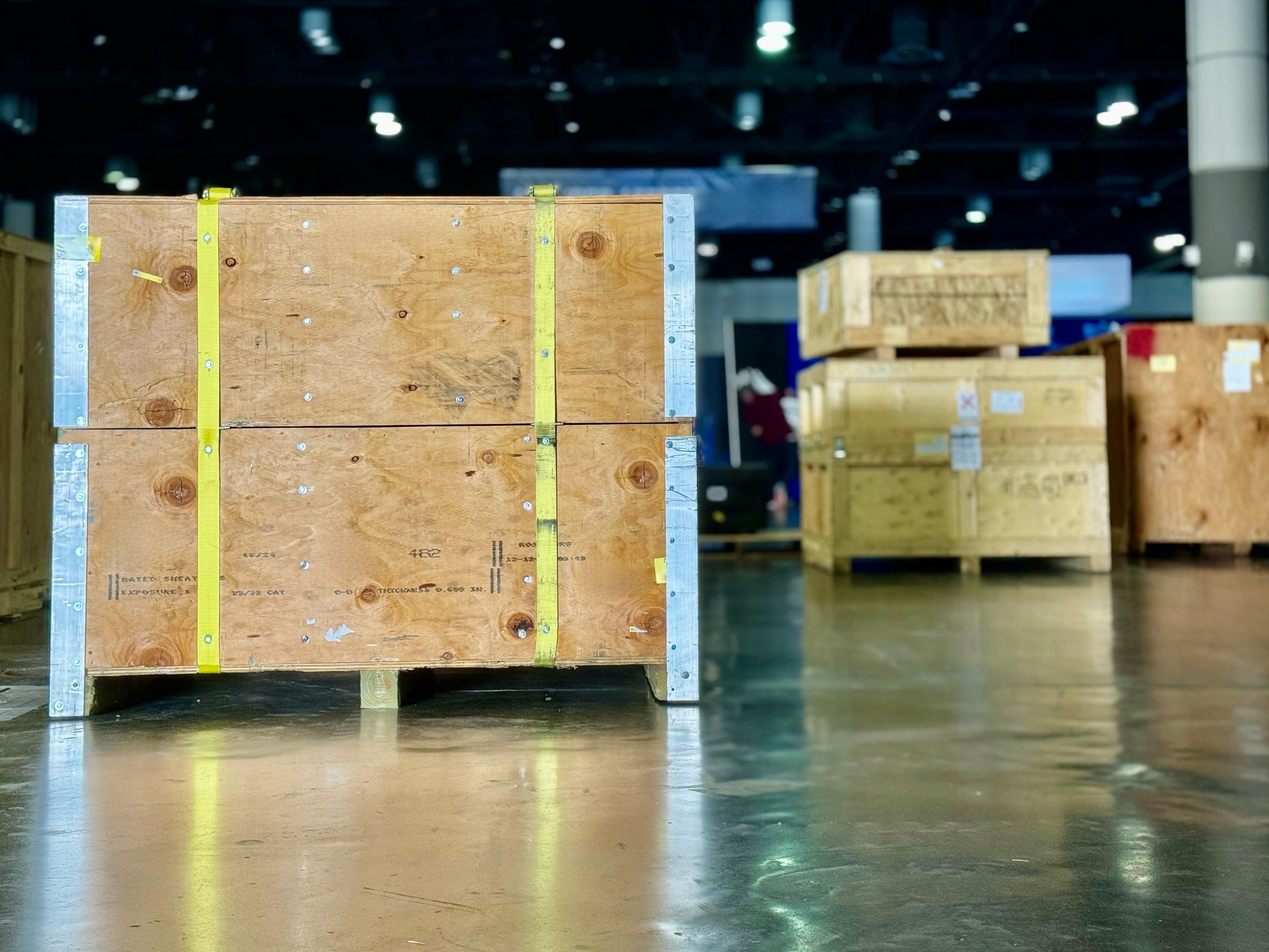 In a dark convention hall, trade show crates sit ready for a fork lift to take them to a booth. The shiny concrete floor reflects the crate against bright direct light. The wood is in good repair.