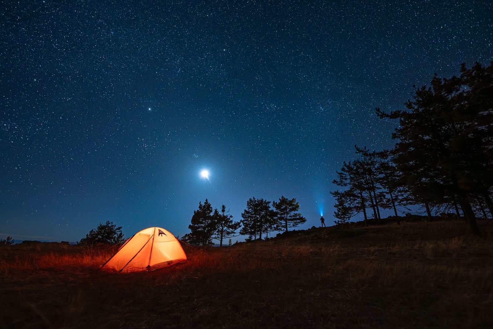 Small silhouette of alone hiker at his illuminated camp tent at starry night. Man with headlight and looking into the distance