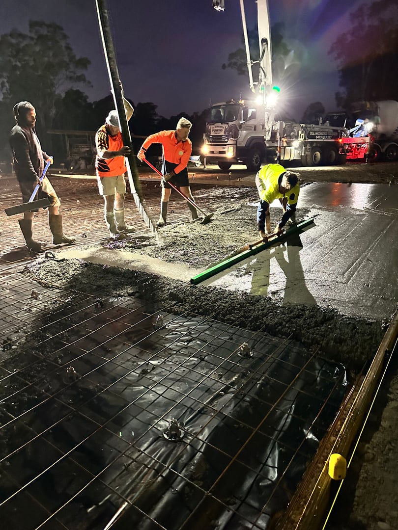 Workers pouring and leveling concrete at a construction site at night, with a concrete mixer truck in the background.