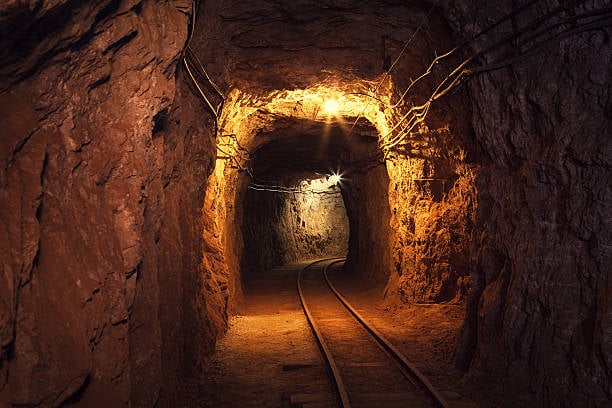 Dimly lit underground mine tunnel with narrow rails and rugged rock walls, illuminated by overhead lights.
