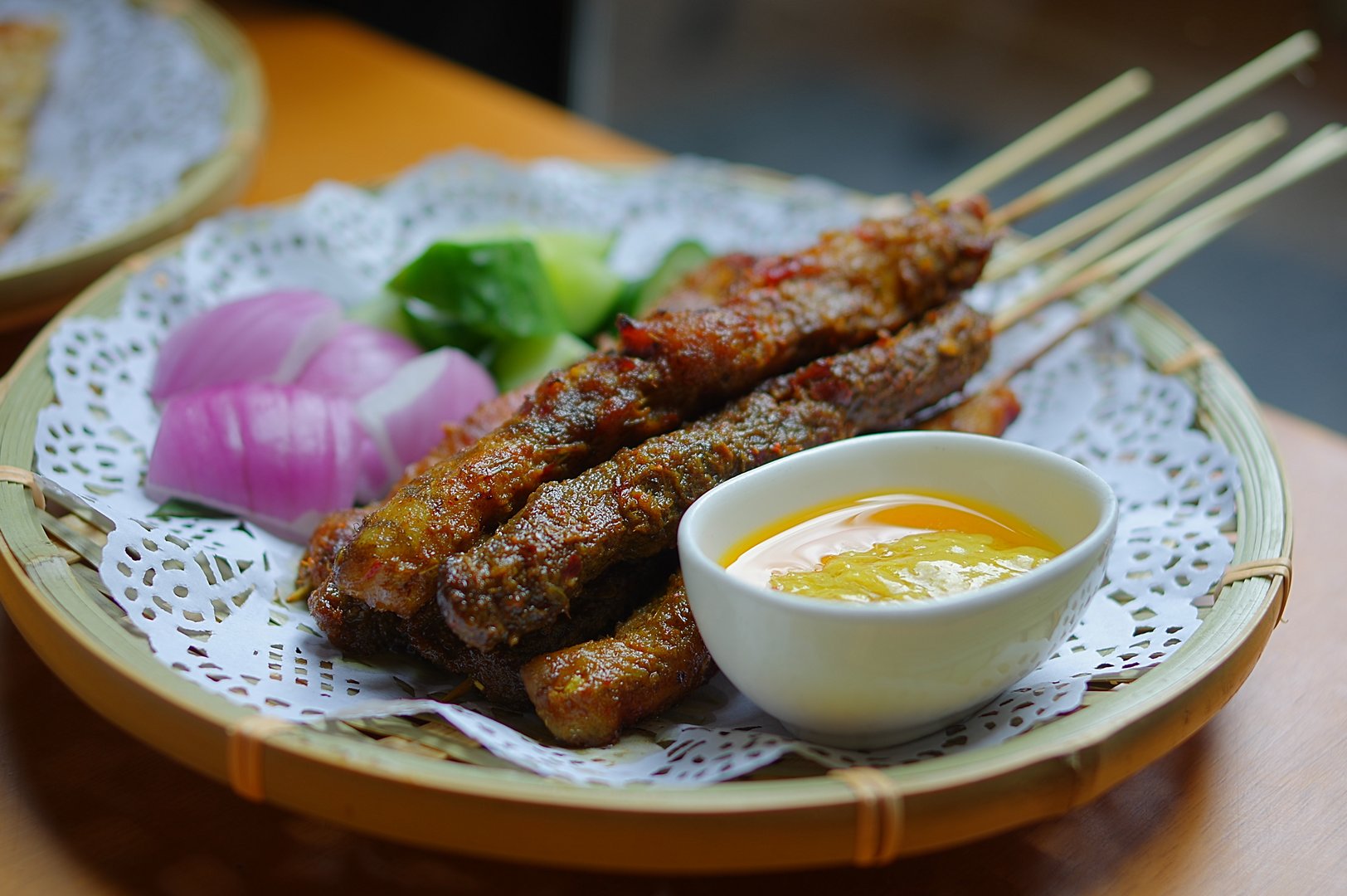Satay beef and chicken meat skewers with sliced onion and cucumber on paper mat and round bamboo plate on table at Southeast Asia cuisine restaurant