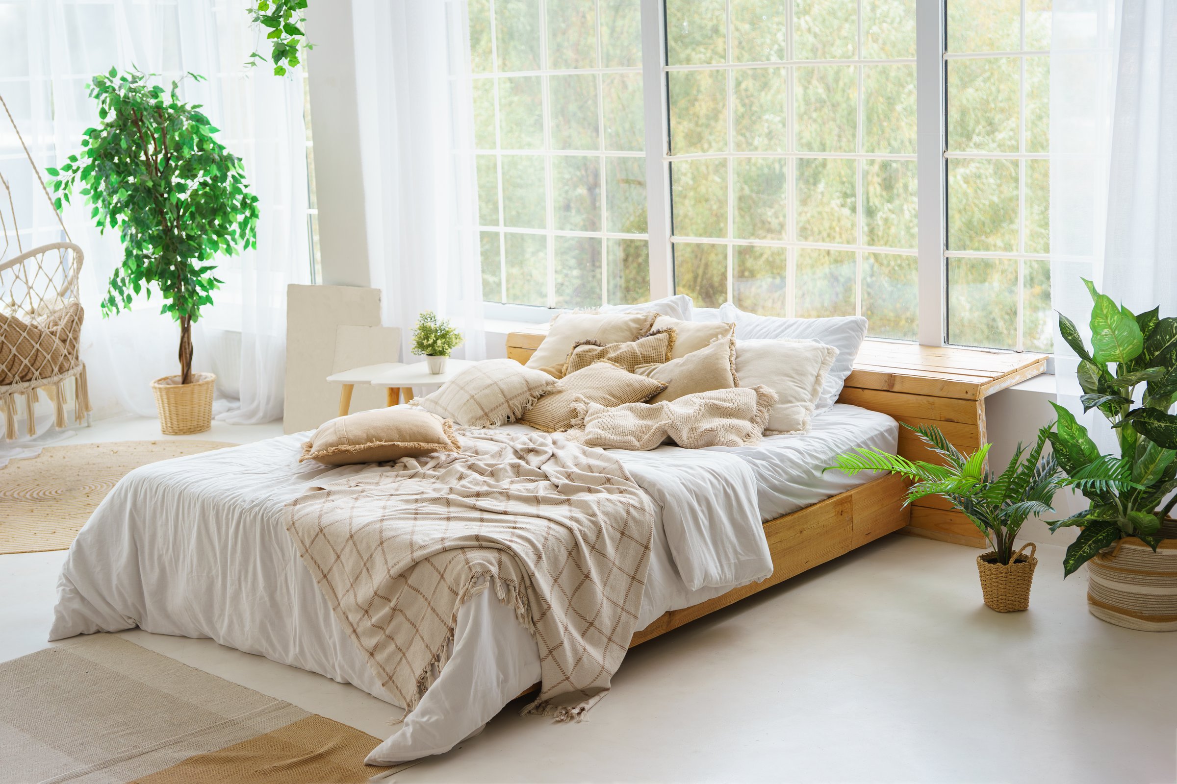Comfortable bed with lots of soft decorative beige pillows in light sunny bedroom with green plants and large window.