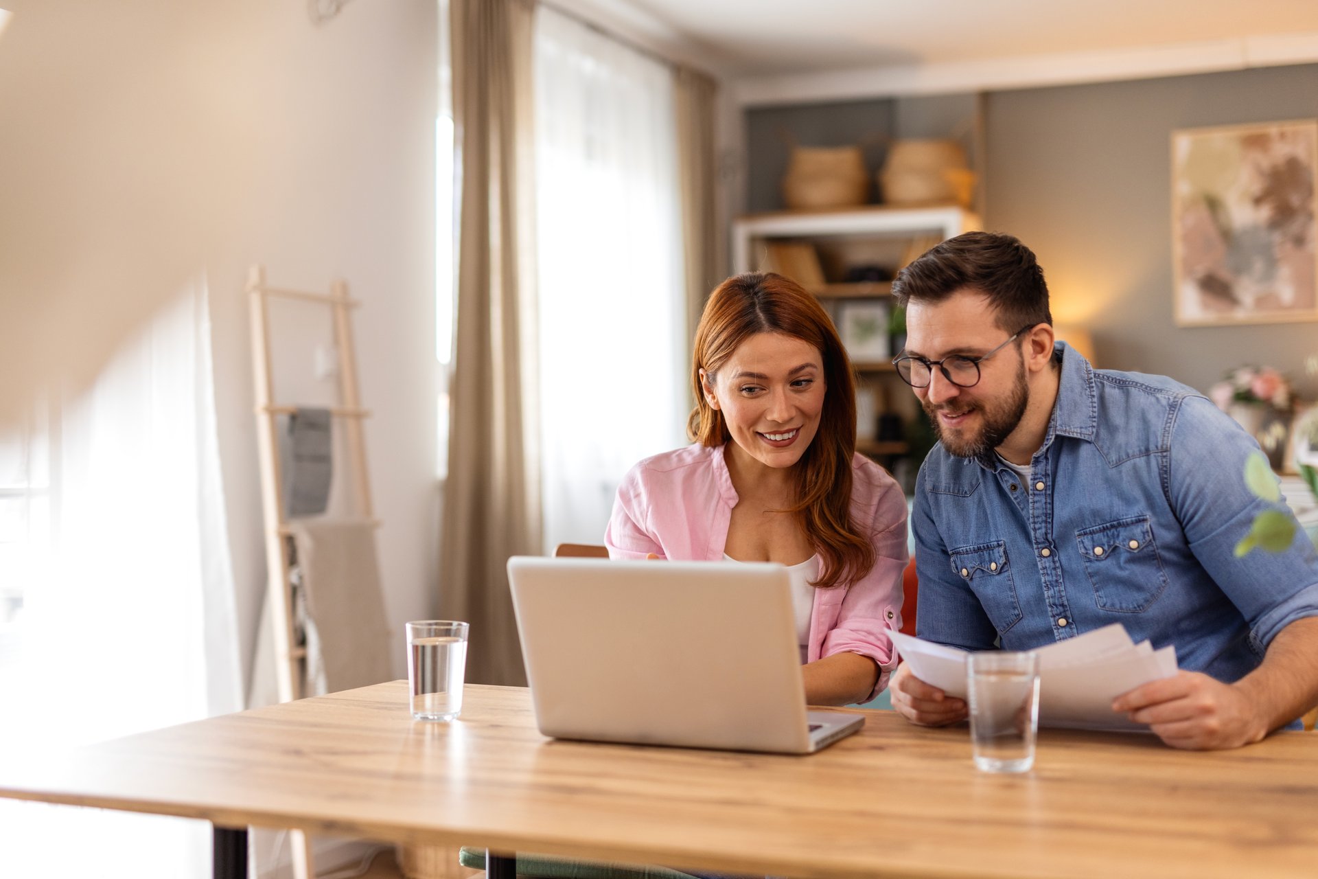 A smiling couple collaborates on financial planning at home, using a laptop and discussing important documents. Their teamwork and organization reflect a positive and balanced lifestyle.