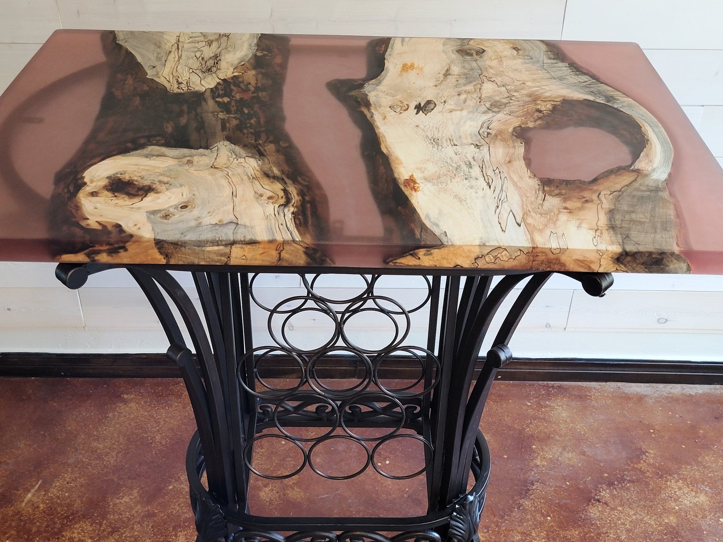 A decorative table with a wooden top featuring rustic wood and resin inlays, supported by a wrought iron wine rack base.