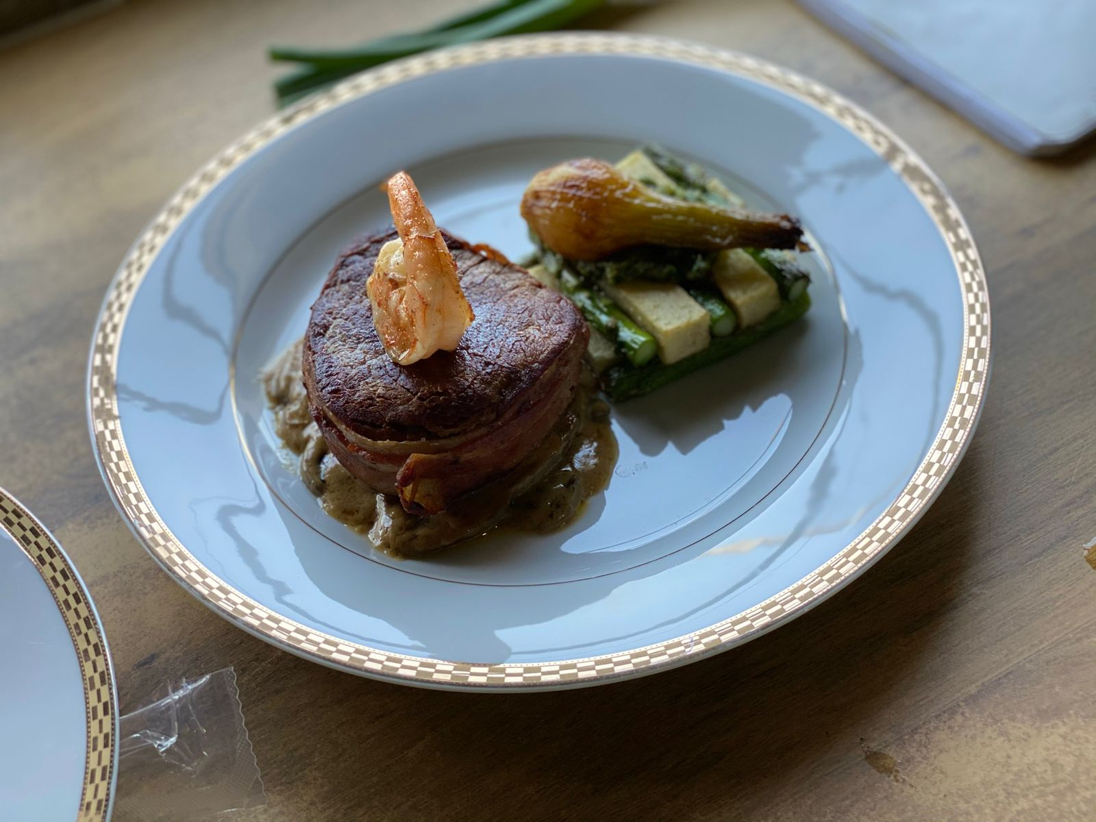 Gourmet dish featuring bacon-wrapped steak, shrimp, and grilled vegetables on an elegant white plate.