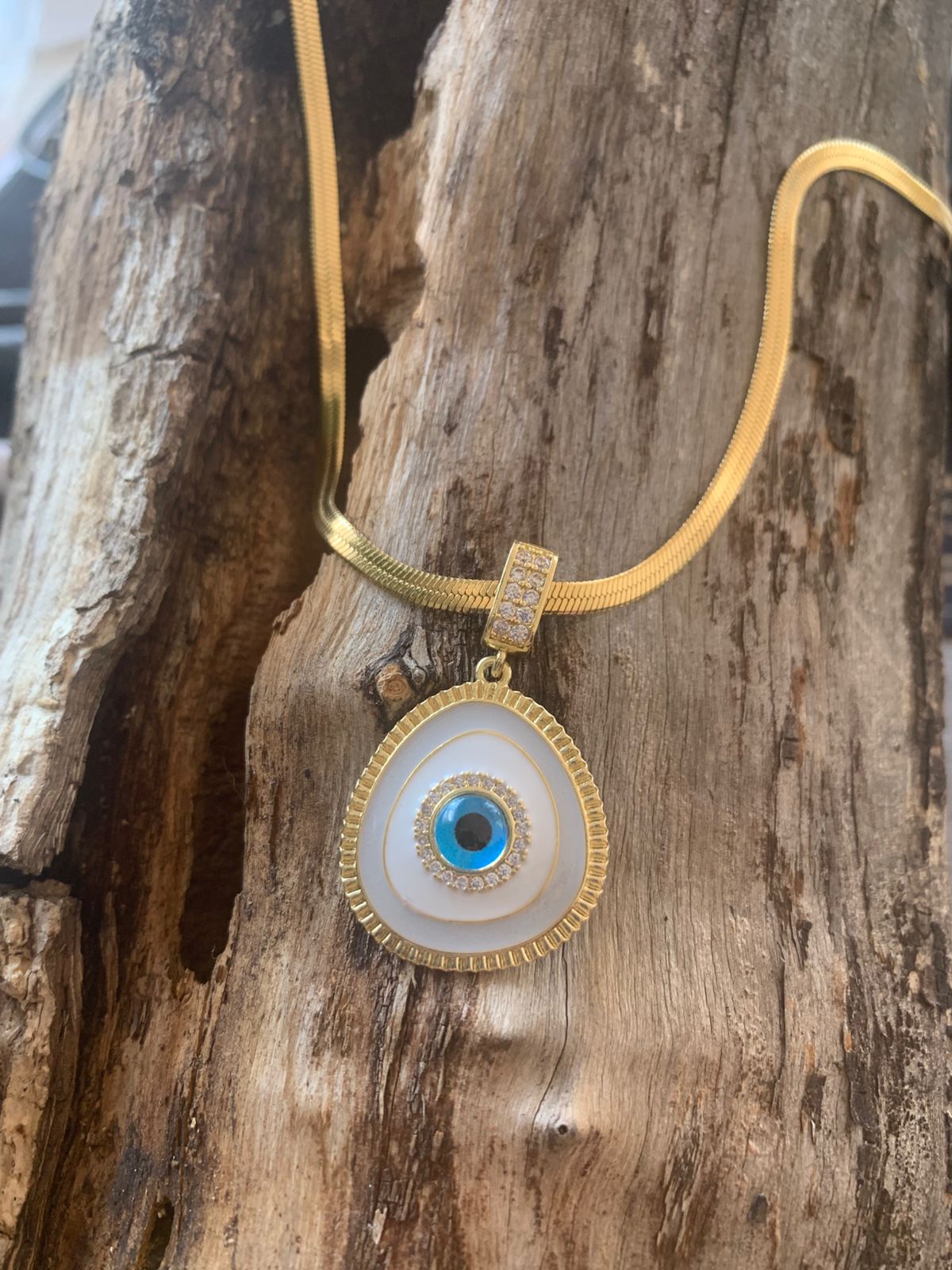 A gold necklace with a large evil eye pendant resting on a textured piece of wood.