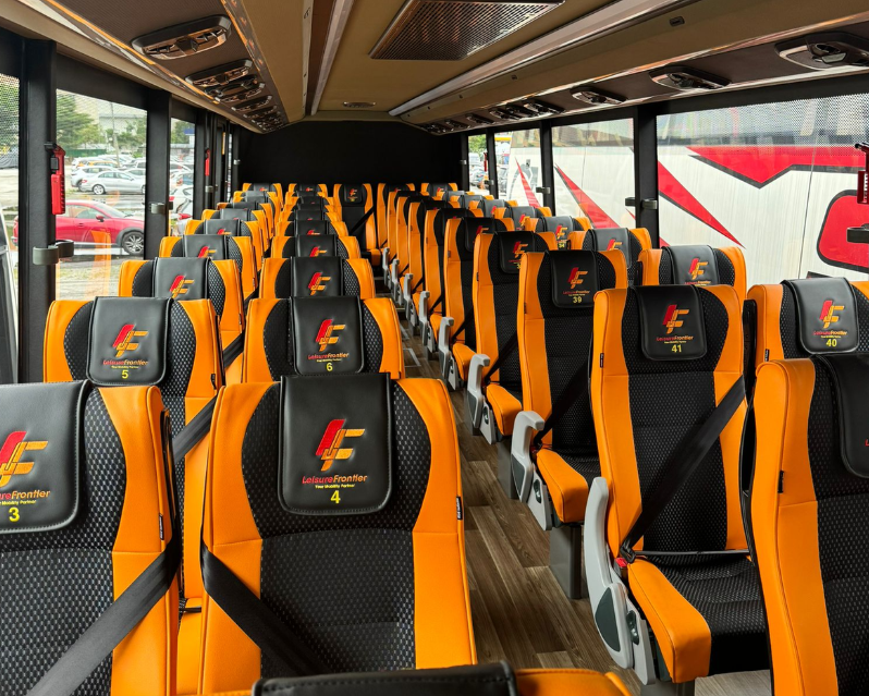 Interior of a luxury bus with orange and black seats featuring seat numbers. Large windows and seat belts visible.