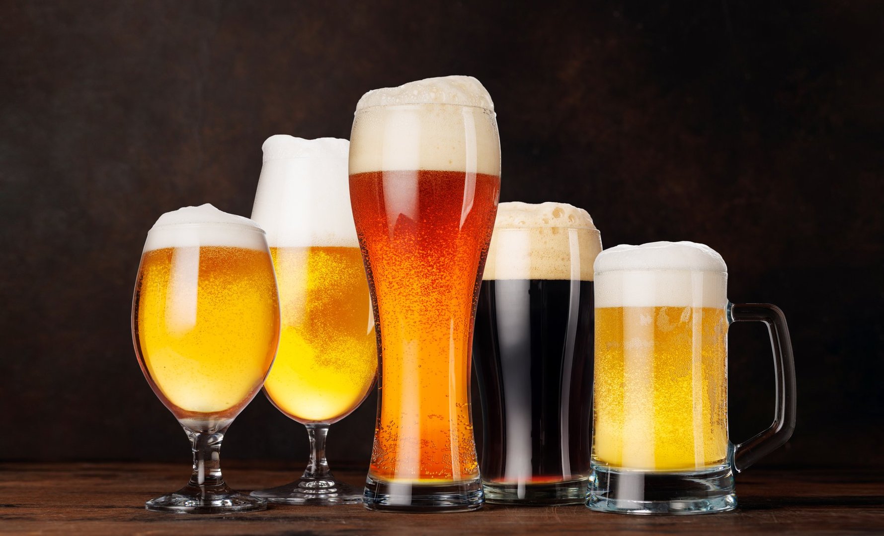 A selection of various beer glasses filled with different types of beer, elegantly arranged on a rustic wooden table against a dark background