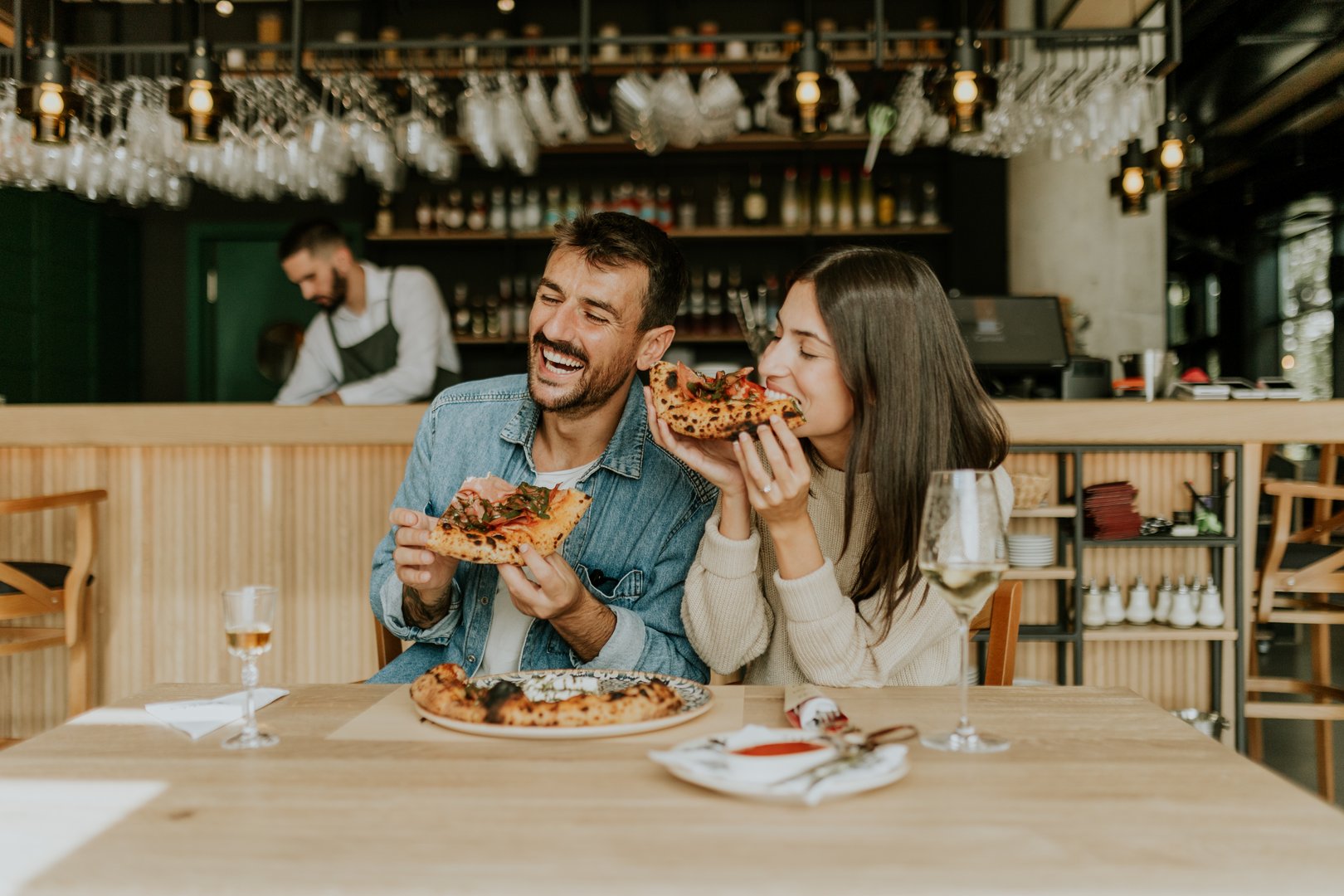 A joyful couple shares a moment at a rustic restaurant, savoring slices of pizza while laughing together and enjoying the vibrant atmosphere around them.
