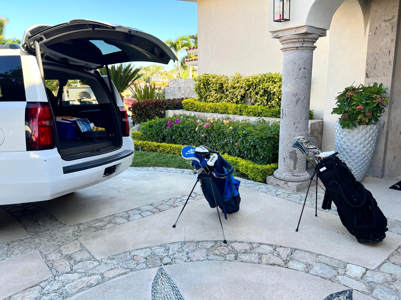 Two golf bags on a driveway, one near an open SUV trunk. A landscaped garden and entrance columns in the background.