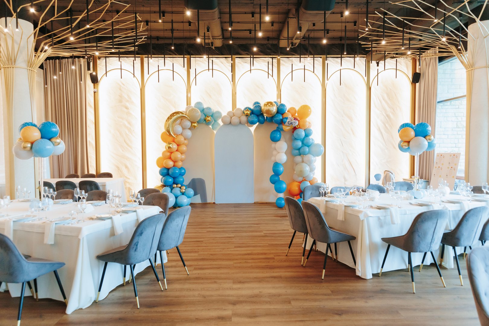 A beautifully decorated event venue featuring a balloon arch in blue, orange, and gold hues, elegant table settings with white tablecloths, and stylish modern chairs.
