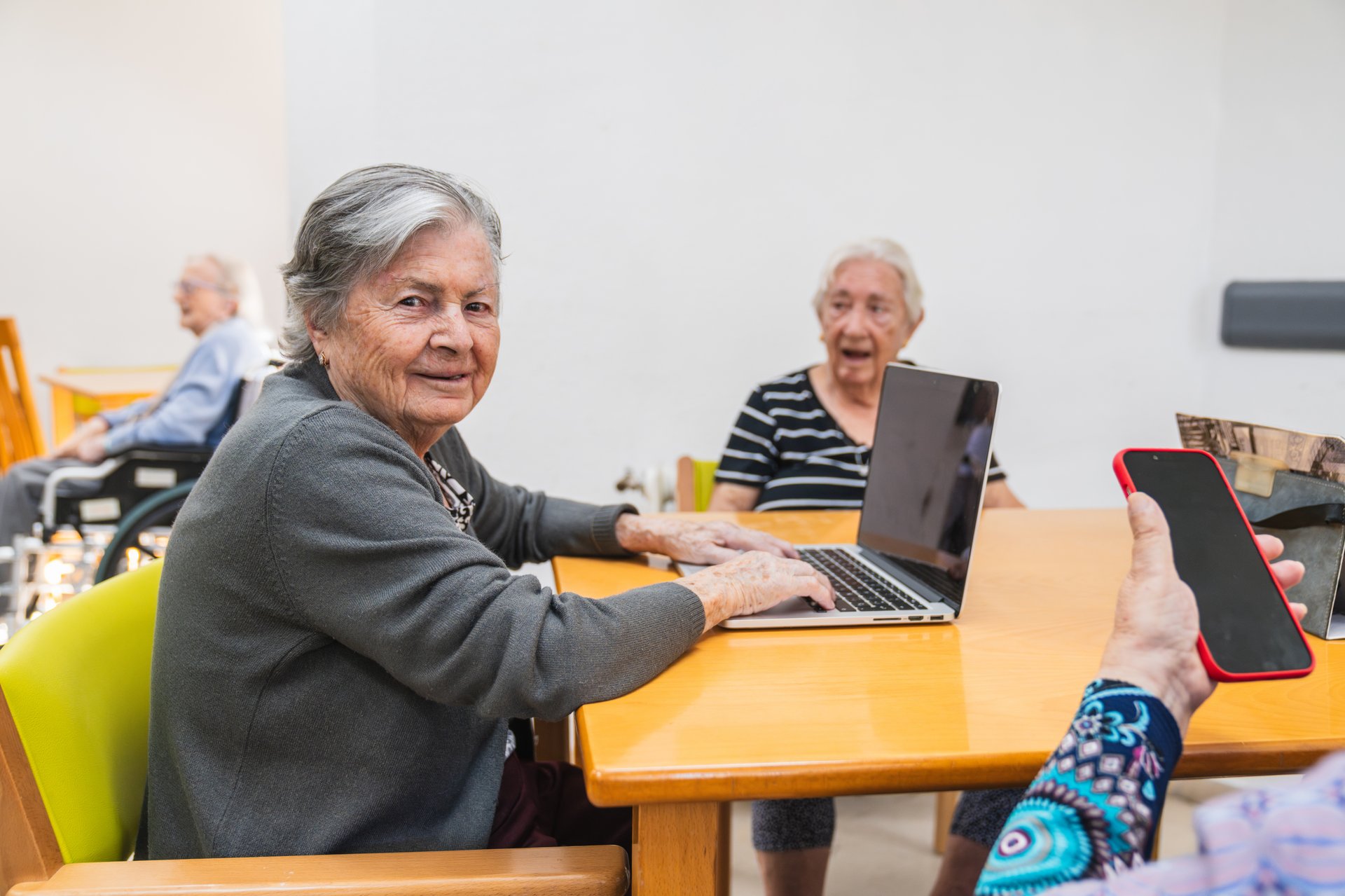 Elderly women in a nursing home are actively using a laptop, showcasing engagement and connection through technology. A caregiver assists with a smartphone