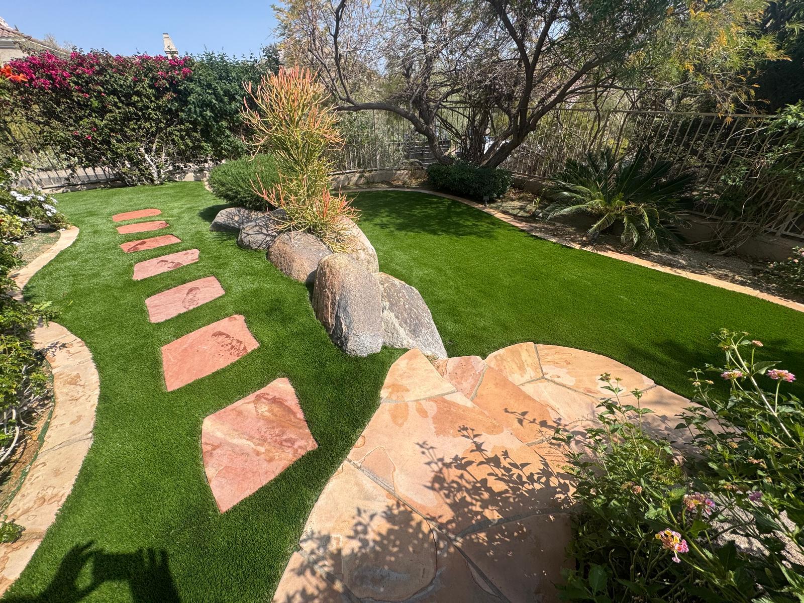 A landscaped garden with a stone path, artificial grass, large rocks, and surrounding greenery and trees.