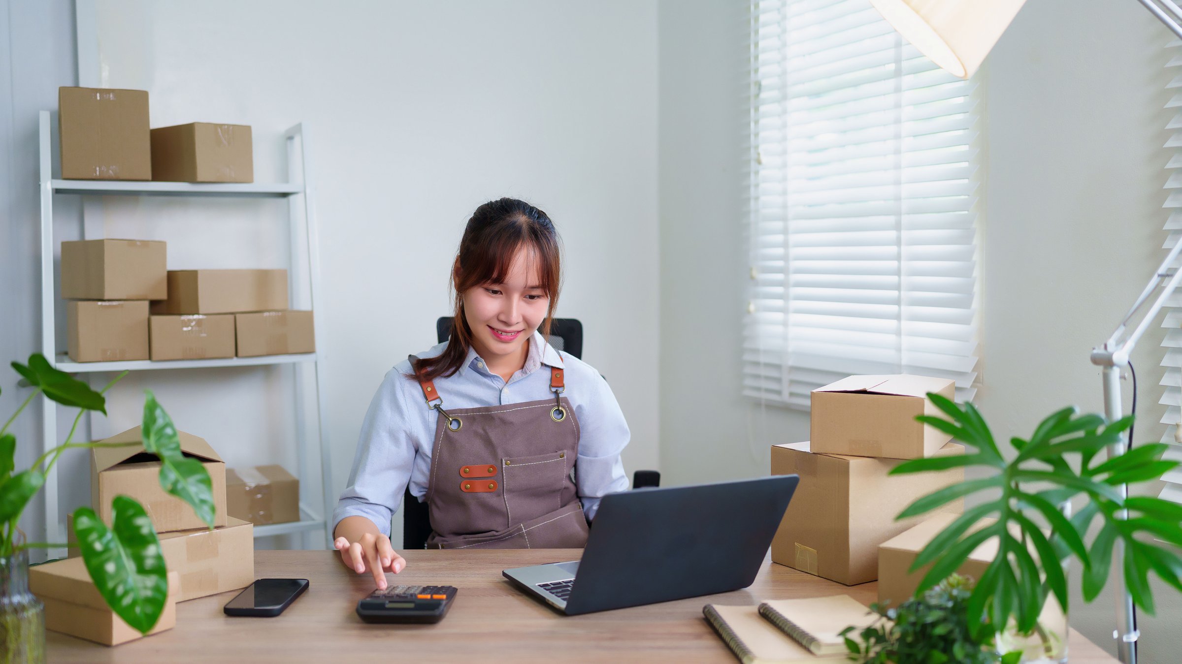 Young Asian women using calculator and laptop to calculate her business expenses for shipping parcels. This is a growing business and she wants to expand her market.