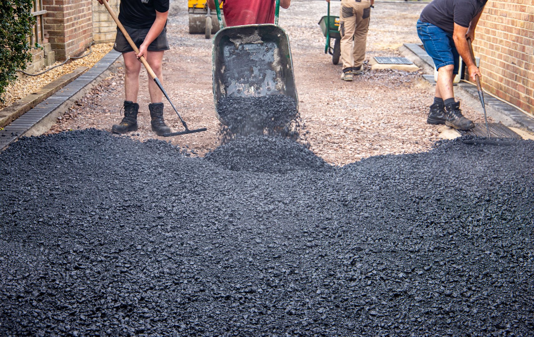 Workers are spreading and leveling asphalt material on a residential driveway in a construction site.