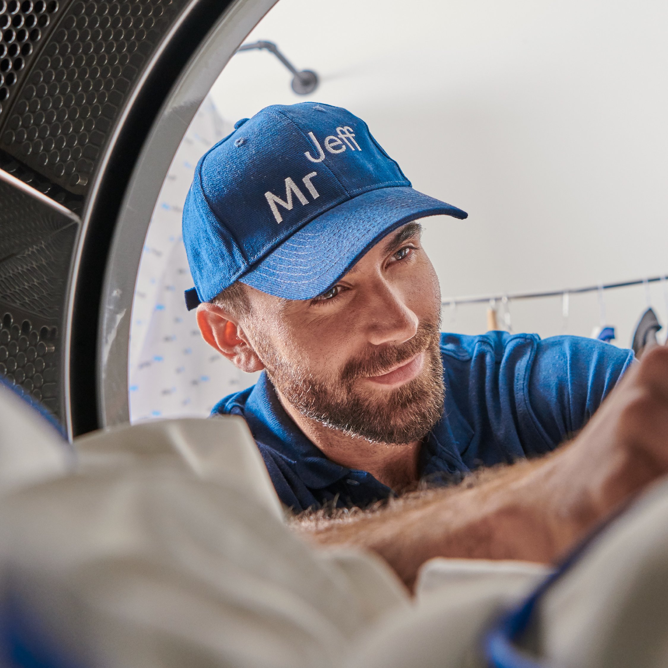 Man with a beard wearing a blue cap and shirt inside a large appliance, possibly a dryer, with laundry items visible.