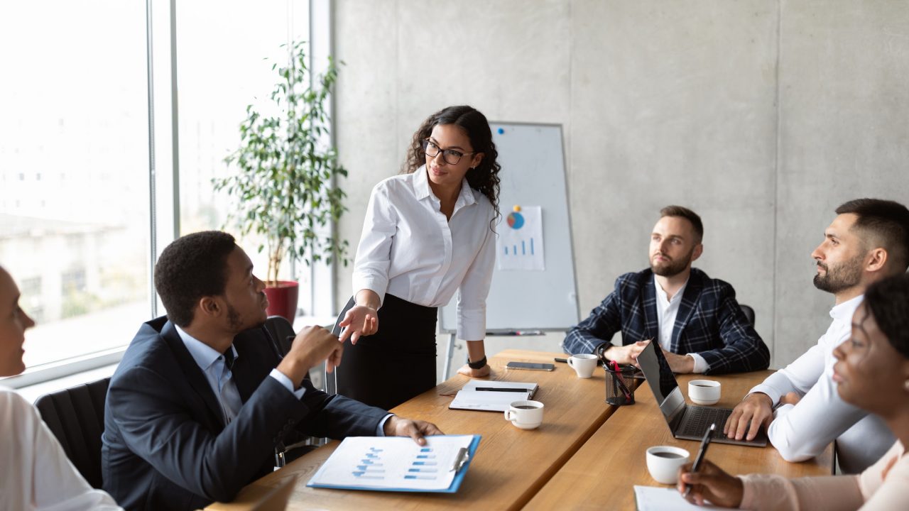 Business meeting with five people discussing around a conference table, charts on a clipboard and whiteboard visible.
