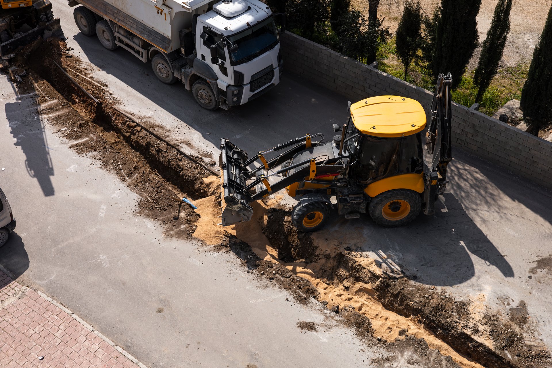 Construction workers are digging a trench alongside a road using a large excavator. Trucks are parked nearby, and the sun illuminates the scene during the day. Dust and soil are visible. Aerial view