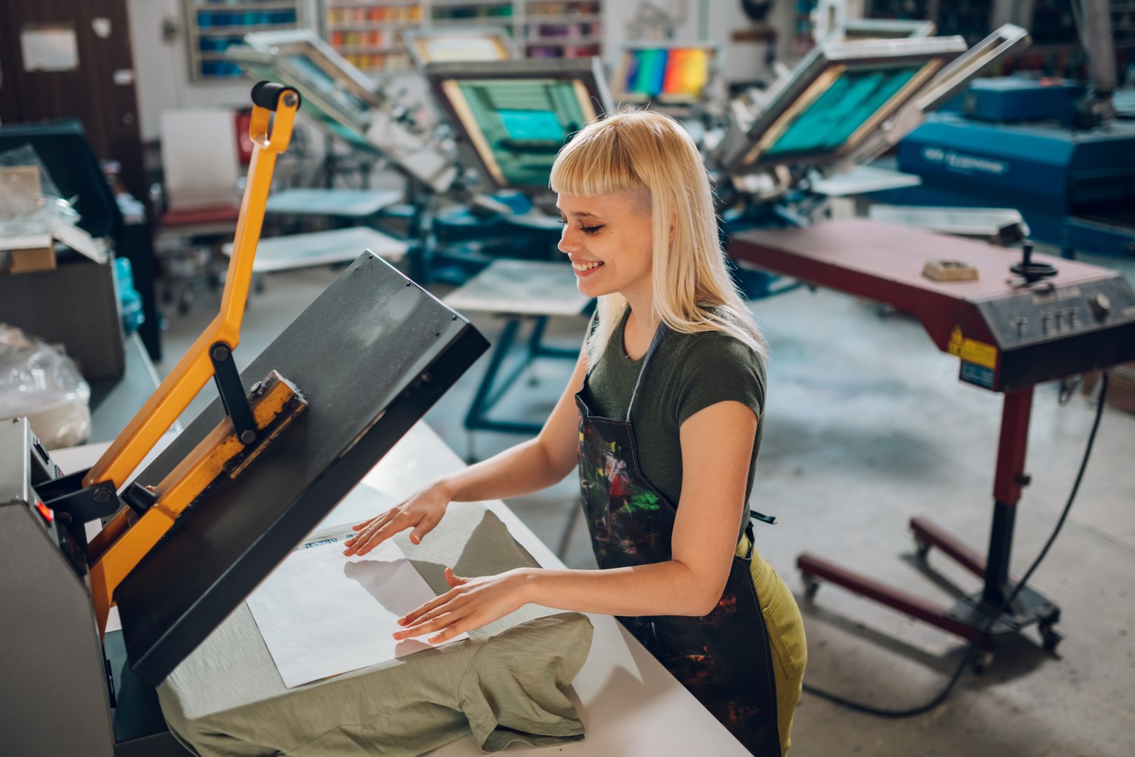 Smiling female printing workshop worker using transfer on a heating press at facility. Happy female graphic technician standing at printing shop and using heating press for transferring. Copy space.