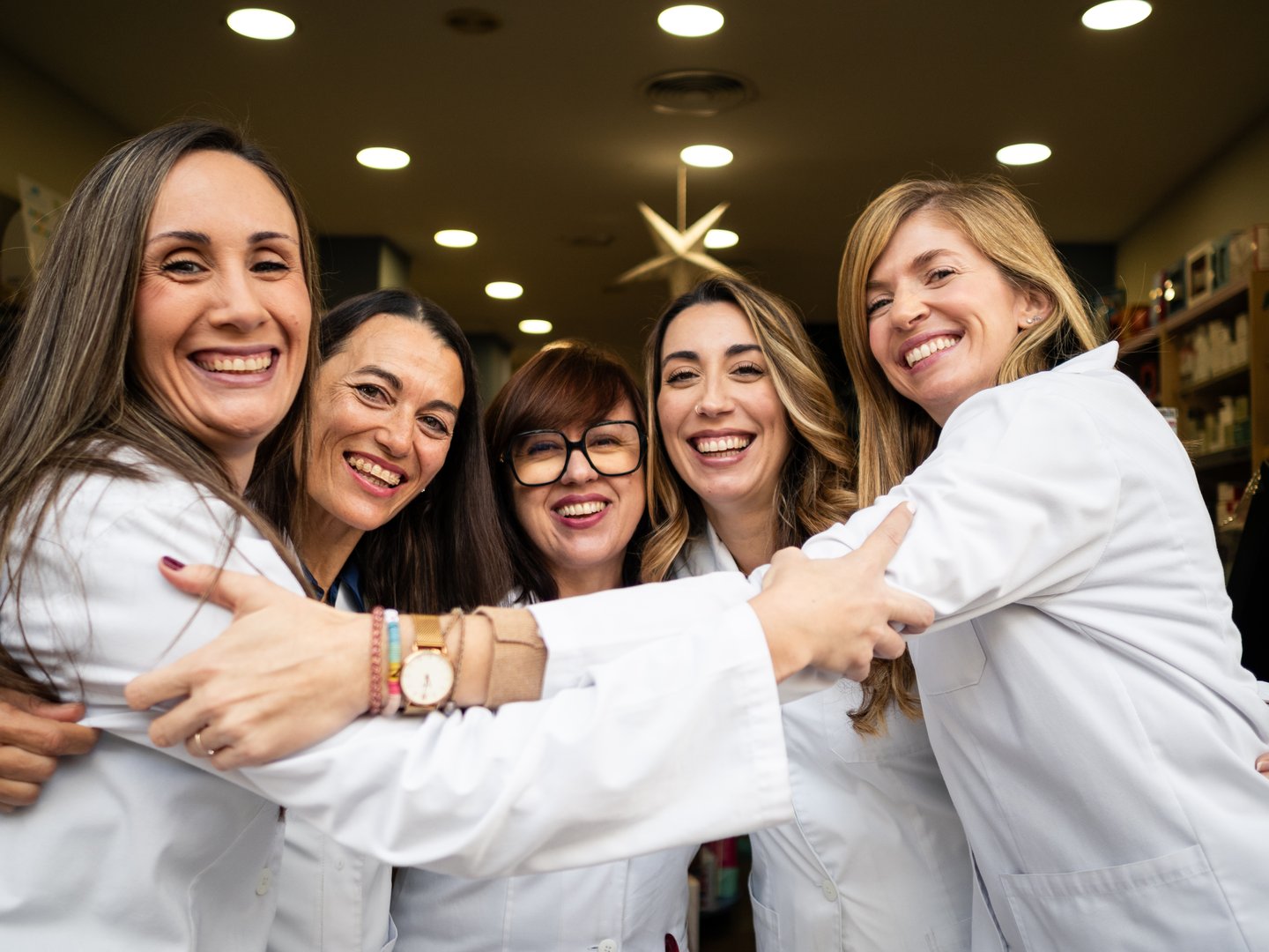 Five women pharmacists smile and embrace each other in a lively pharmacy. They showcase camaraderie and professionalism in their white coats, reflecting a thriving community environment.