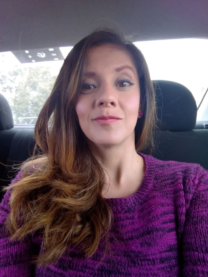 Woman with long hair wearing a purple sweater, seated in a car, smiling at the camera.