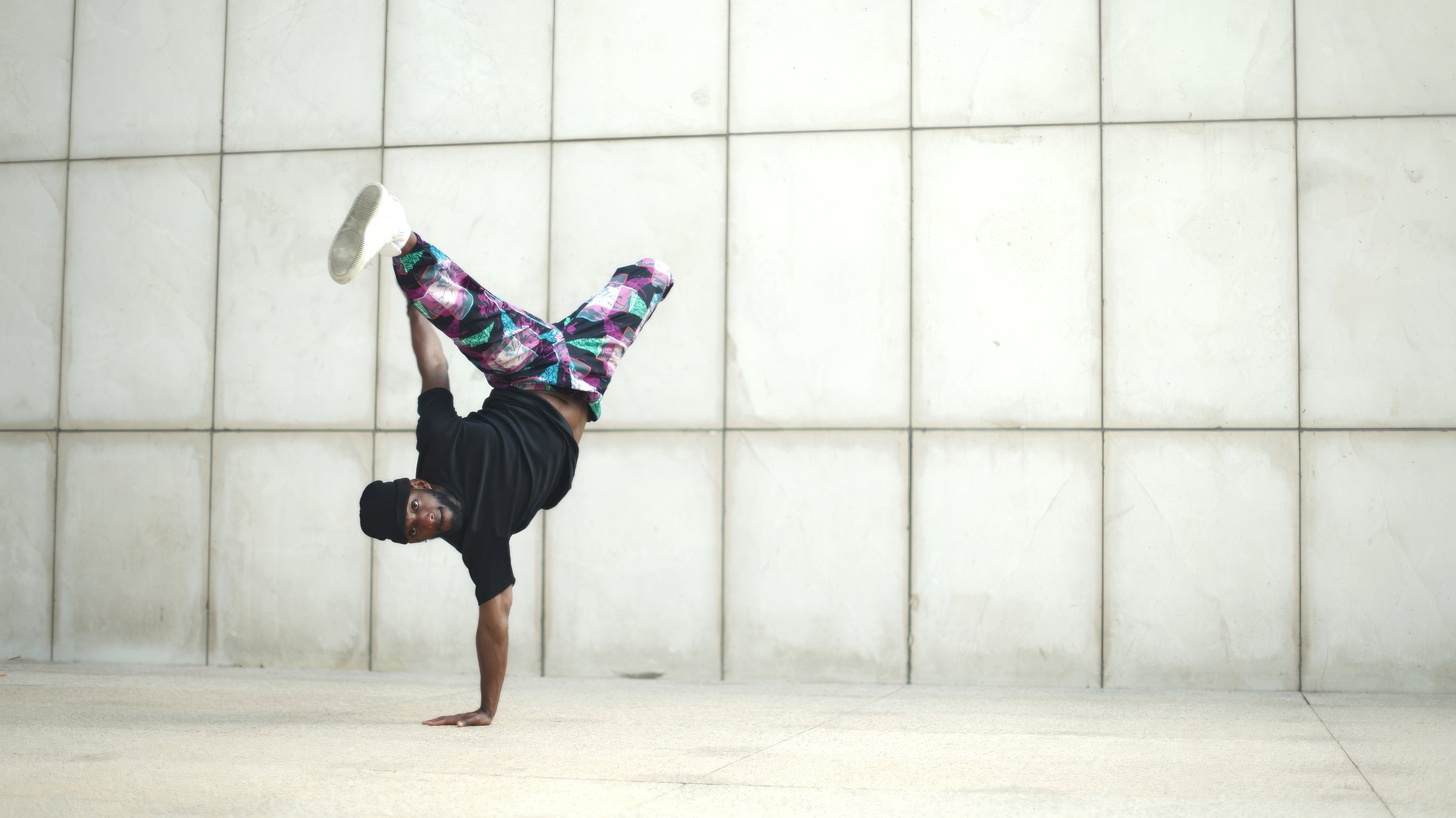 Young breakdancer balancing on his hands with his legs spread, performing in front of a white tiled wall