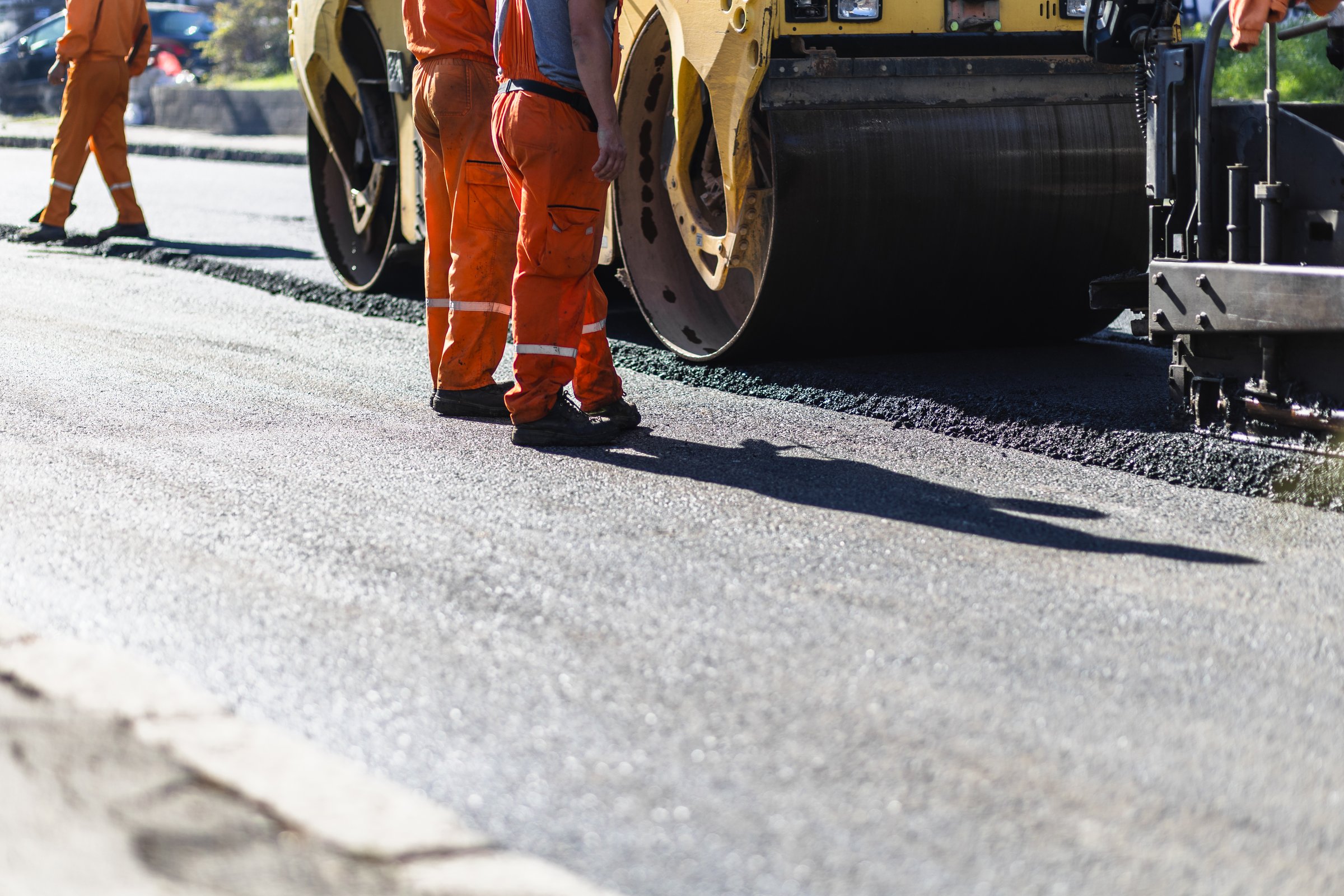 A team of construction workers in bright orange uniforms is overseeing the laying of asphalt. Heavy machinery is actively compacting the new road surface under clear skies.